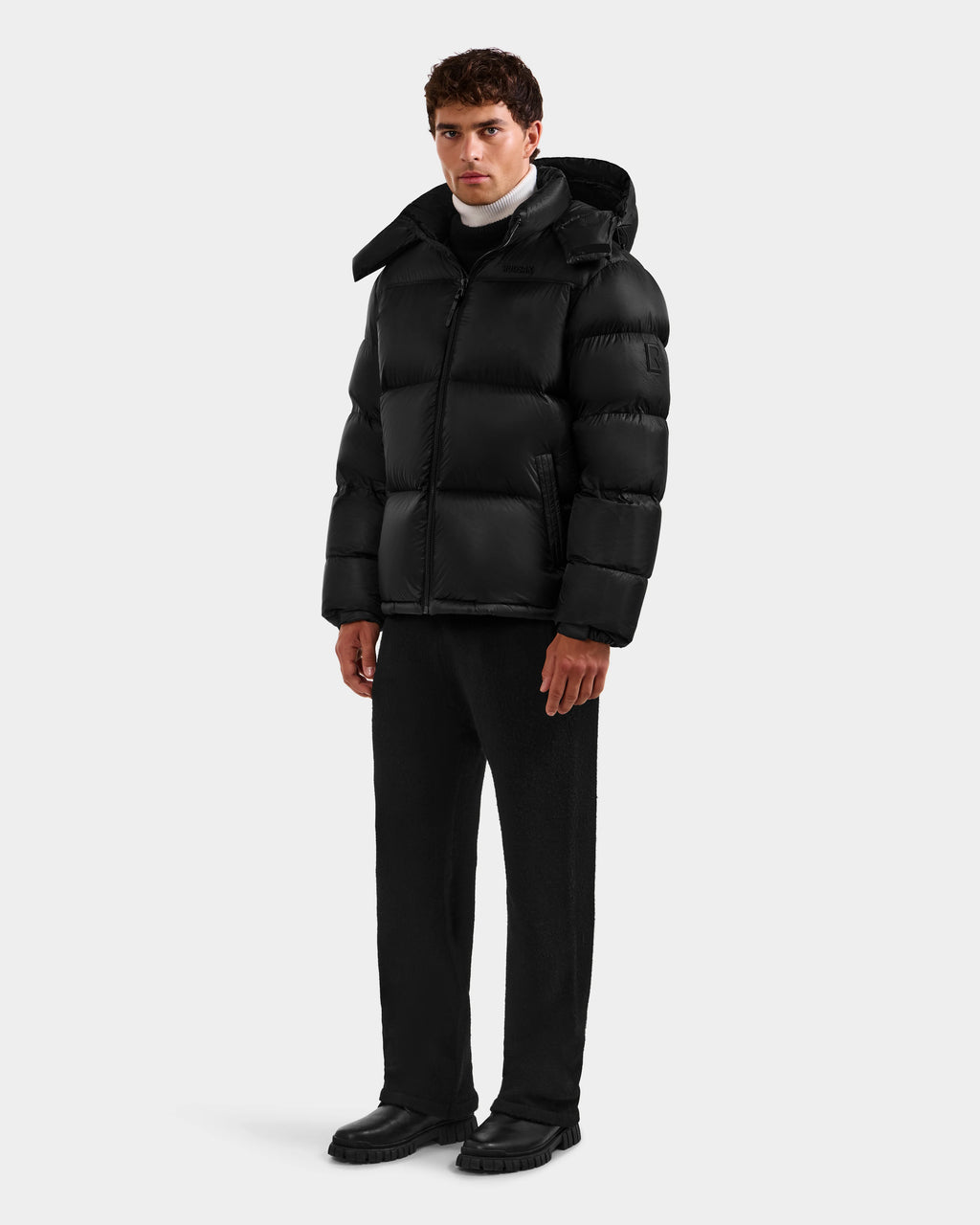A man faces forward wearing the Rudsak STEFANO - 6125574 Black men’s down puffer jacket, paired with black pants, black shoes, and a white turtleneck against a plain light background.