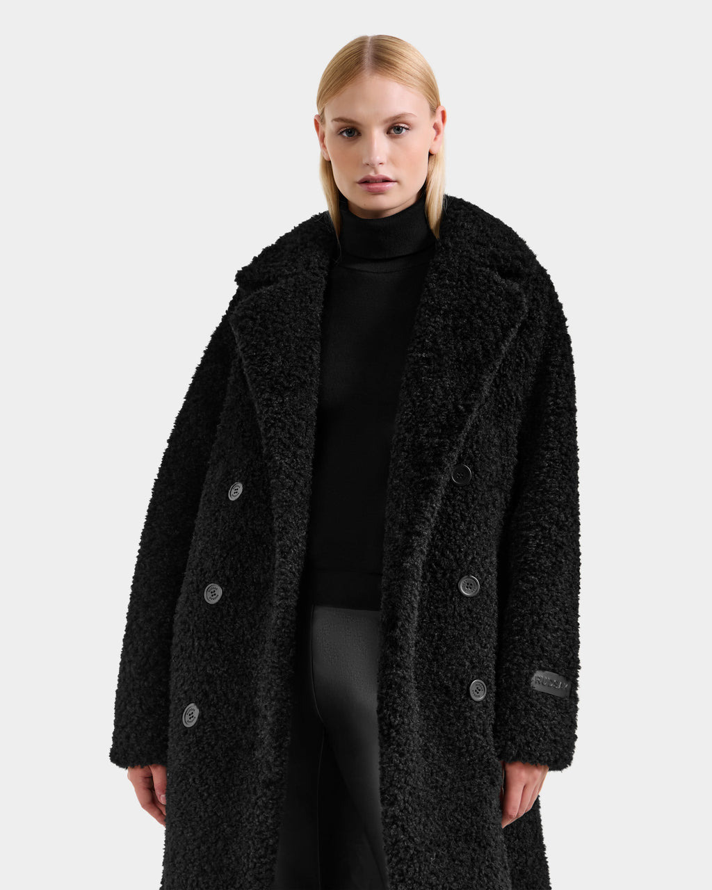 A woman with straight blonde hair models the Rudsak TINA - 8125968 Black faux fur teddy trench coat with removable hooded bib over a black turtleneck and pants, standing against a plain light gray background.