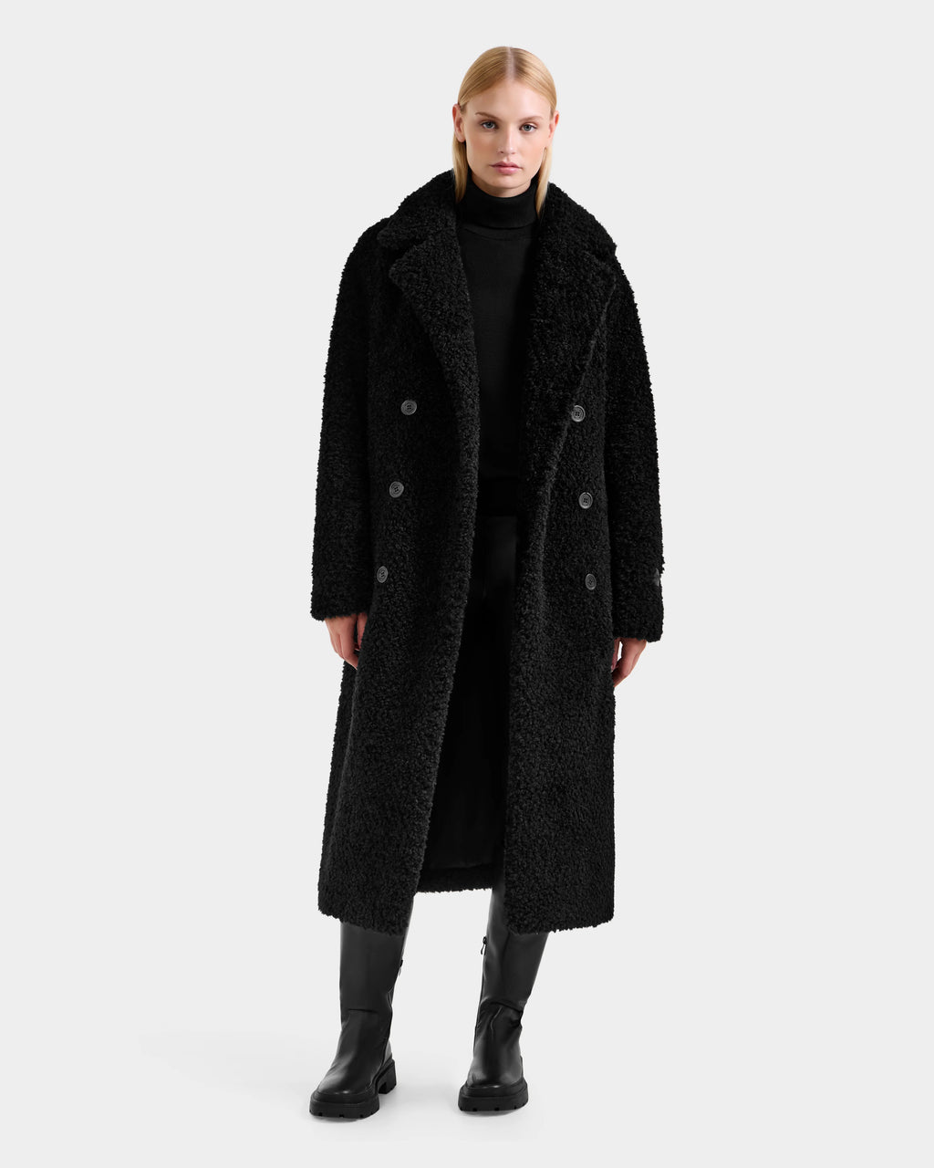 A woman stands facing forward in the Rudsak TINA - 8125968 Black faux fur teddy trench coat with a removable hooded bib, styled over a black turtleneck, pants, and boots against a plain white background.