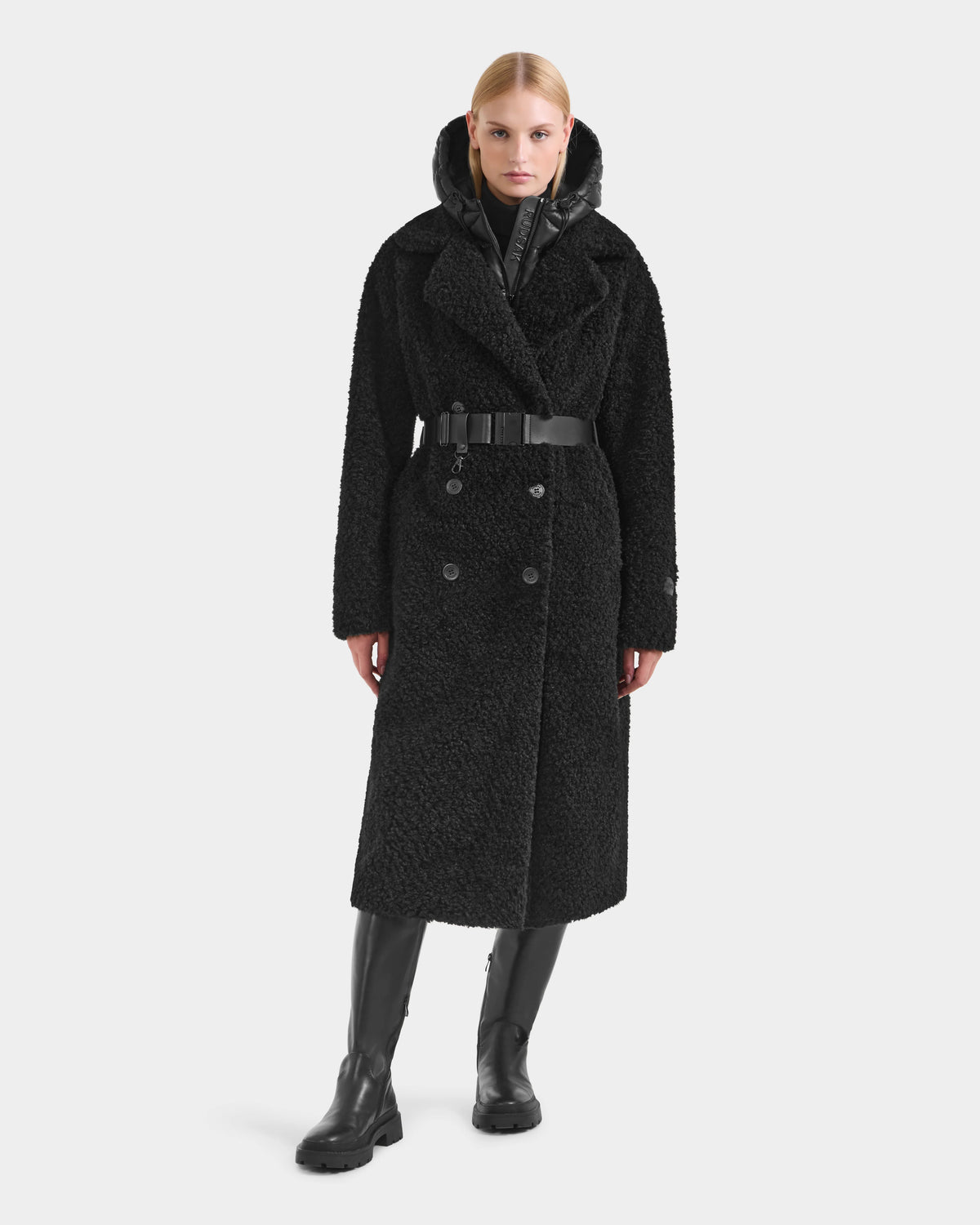 A woman with blonde hair styled back and a neutral expression faces forward, wearing the Rudsak TINA - 8125968 Black textured faux fur teddy trench coat with a wide belt, black boots, and a high-collared inner layer.