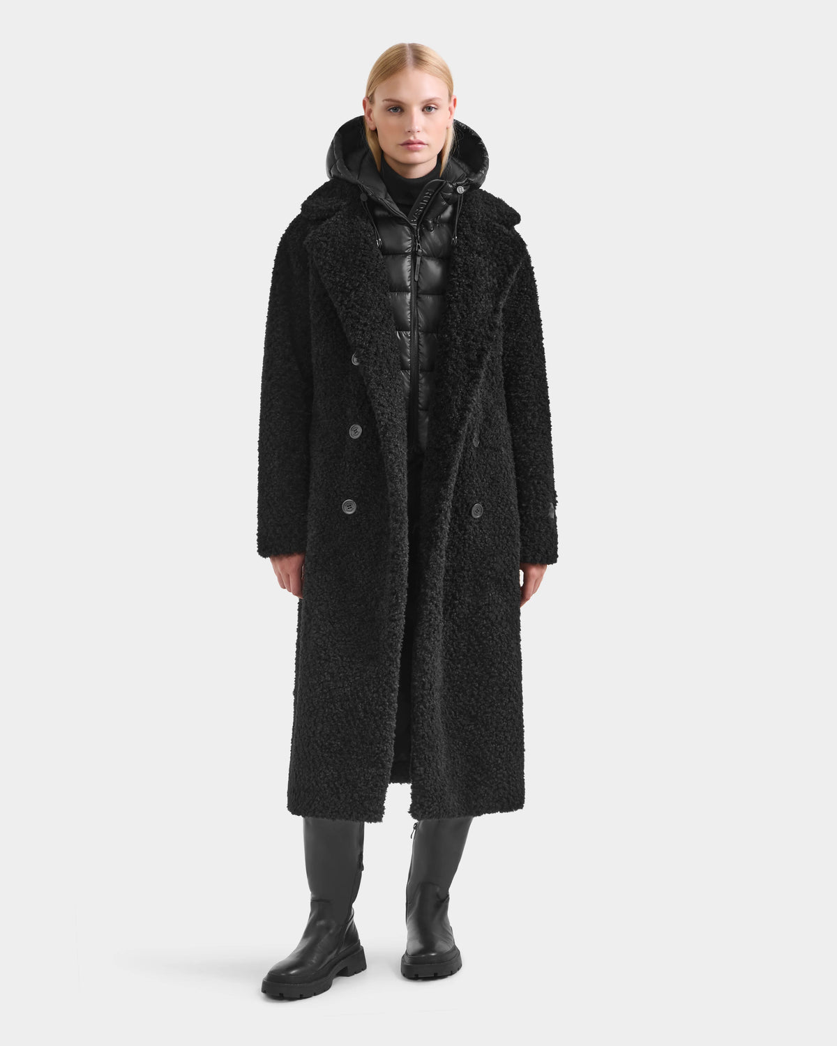 A person stands facing forward, wearing the Rudsak TINA - 8125968 Black faux fur teddy trench coat over a black puffer jacket with removable hooded bib, paired with black pants and boots against a plain light background.