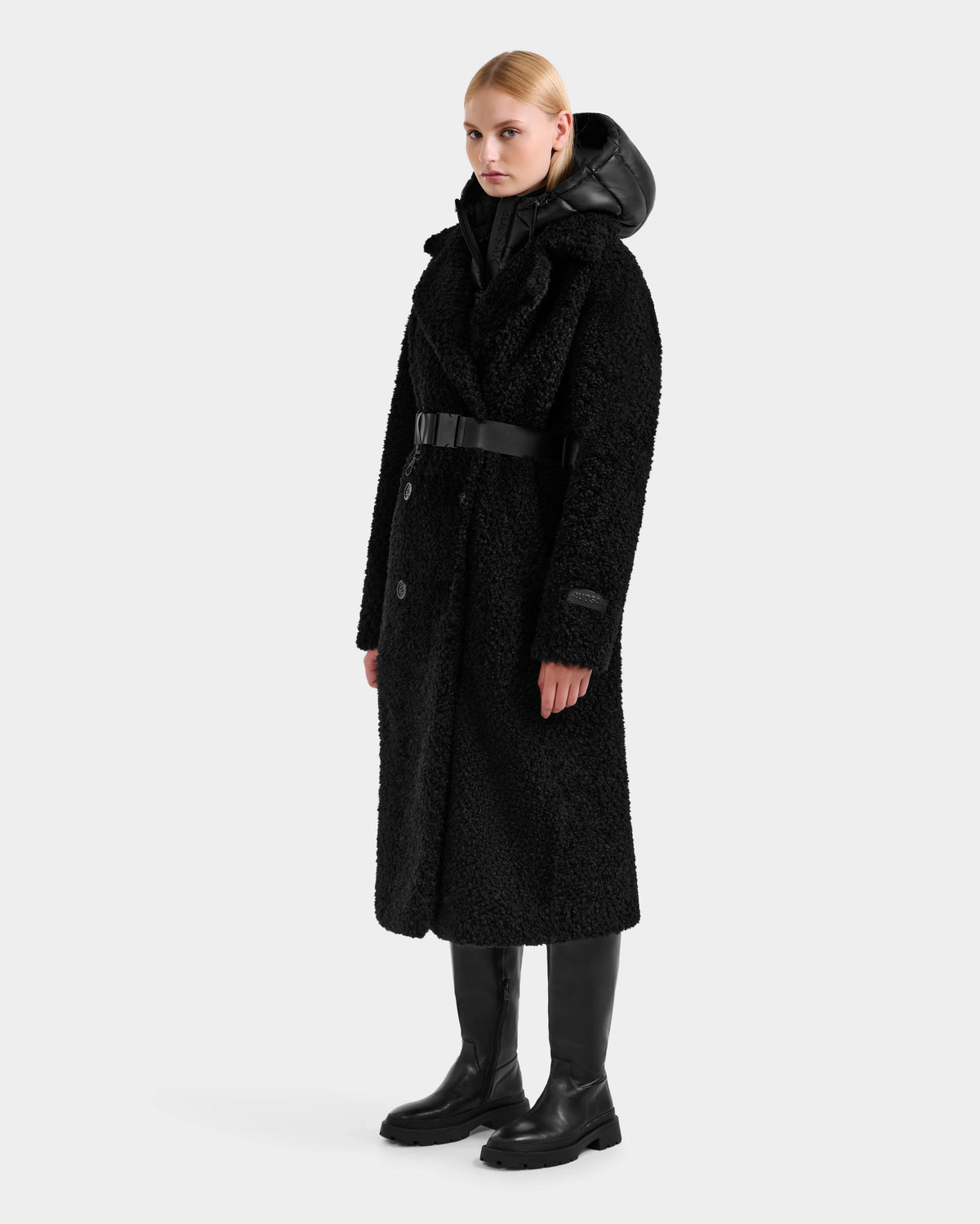A woman models the Rudsak TINA - 8125968 Black faux fur teddy trench coat with a wide collar, black belt, and knee-high boots. She layers it over a removable hooded bib against a plain light grey background.