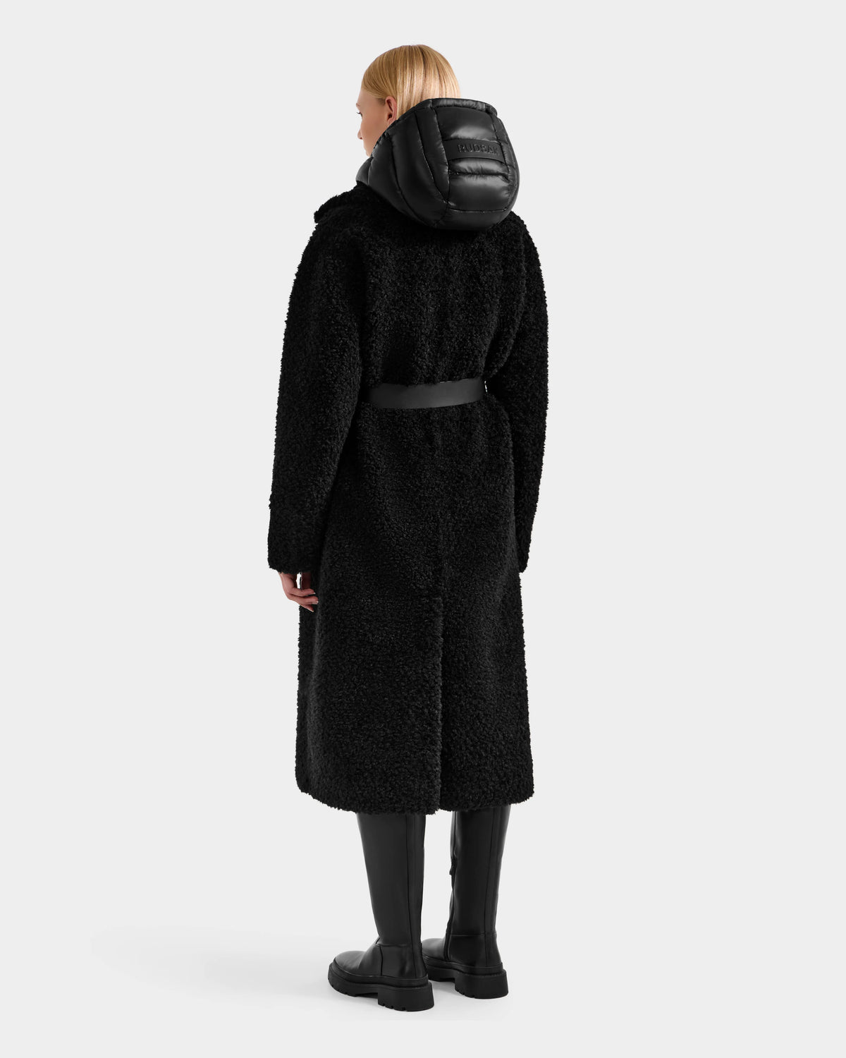 A woman faces away, wearing the Rudsak TINA - 8125968 Black faux fur teddy trench coat with a belt, styled with a black puffer hood and knee-high boots against a plain light background.