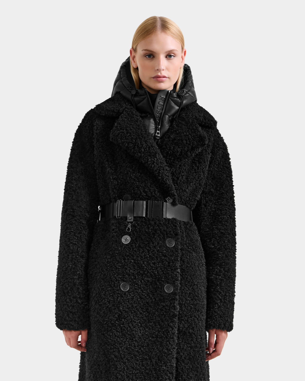 A person faces forward in a textured black double-breasted Rudsak TINA - 8125968 coat with large lapels, removable hooded bib, belted waist, and a black puffer hood. The background is plain light gray.