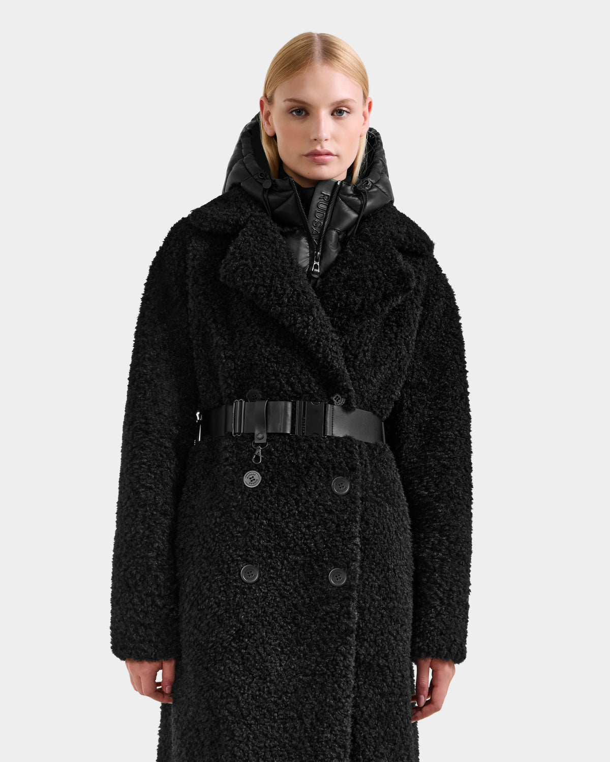 A person faces forward in a textured black double-breasted Rudsak TINA - 8125968 coat with large lapels, removable hooded bib, belted waist, and a black puffer hood. The background is plain light gray.