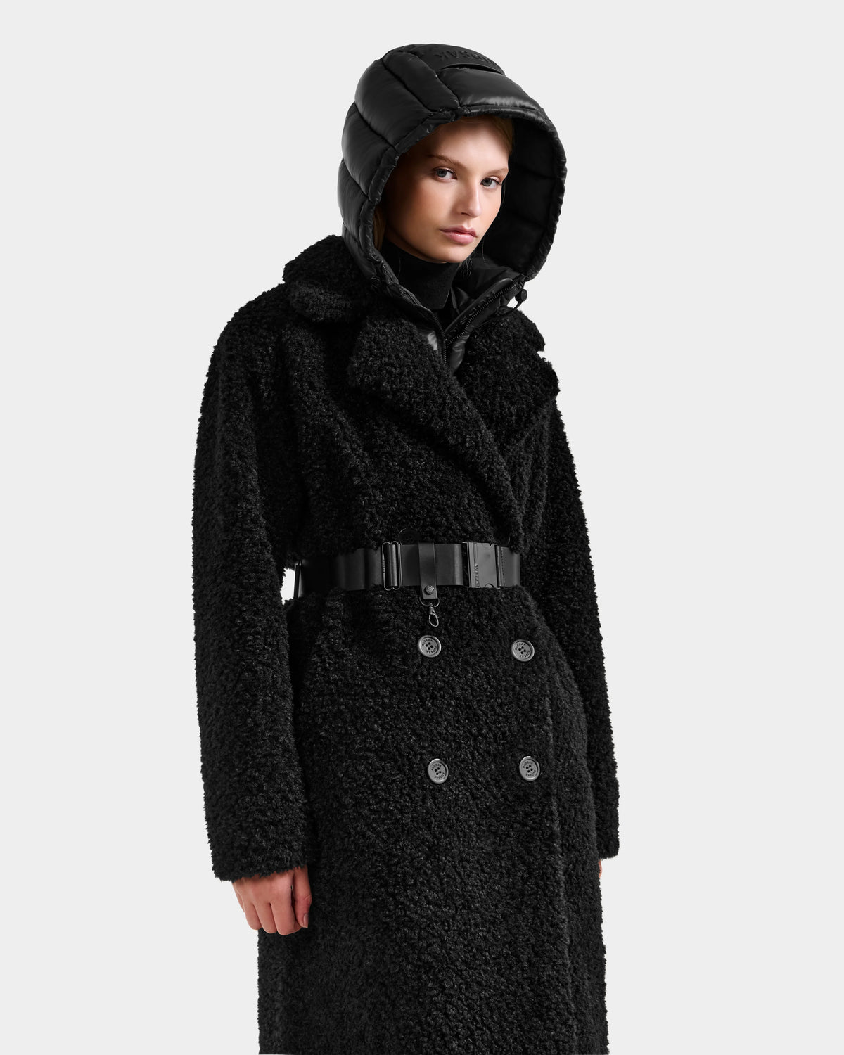 A person wearing the Rudsak TINA - 8125968 Black faux fur teddy trench coat with a wide belt and shiny removable hooded bib, standing against a plain light background.