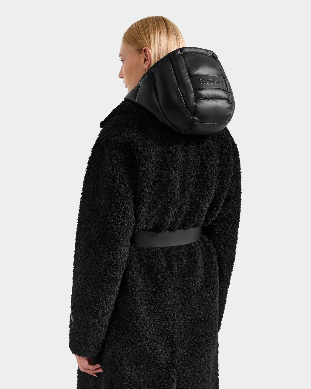 A woman with straight blonde hair, seen from the back, wears the Rudsak TINA - 8125968 Black textured faux fur teddy trench coat with a belted waist and shiny removable hooded bib. The soft, quilted coat stands out against a plain light gray background.