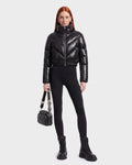 A person with long red hair wears the Rudsak ANSLIE C - 8126713 Black, a shiny, fashion-forward puffer jacket made with RDS-certified down, paired with black leggings, lace-up black boots, and holding a small black handbag against a plain light background.