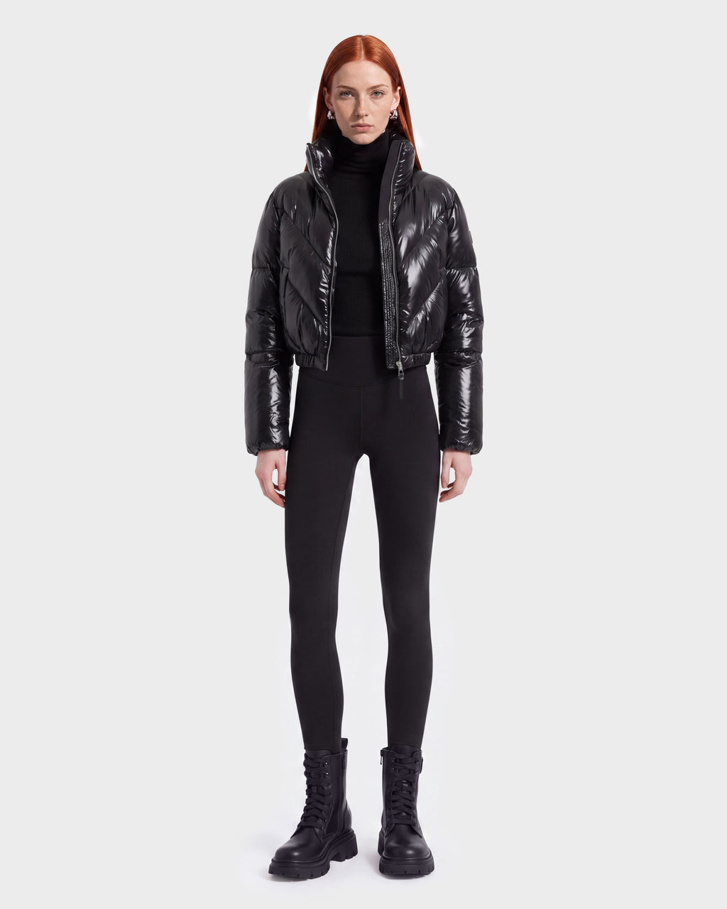 A person with long red hair wears the Rudsak ANSLIE C - 8126713 Black puffer jacket, paired with a black turtleneck, black leggings, and black lace-up boots, standing against a plain white background.