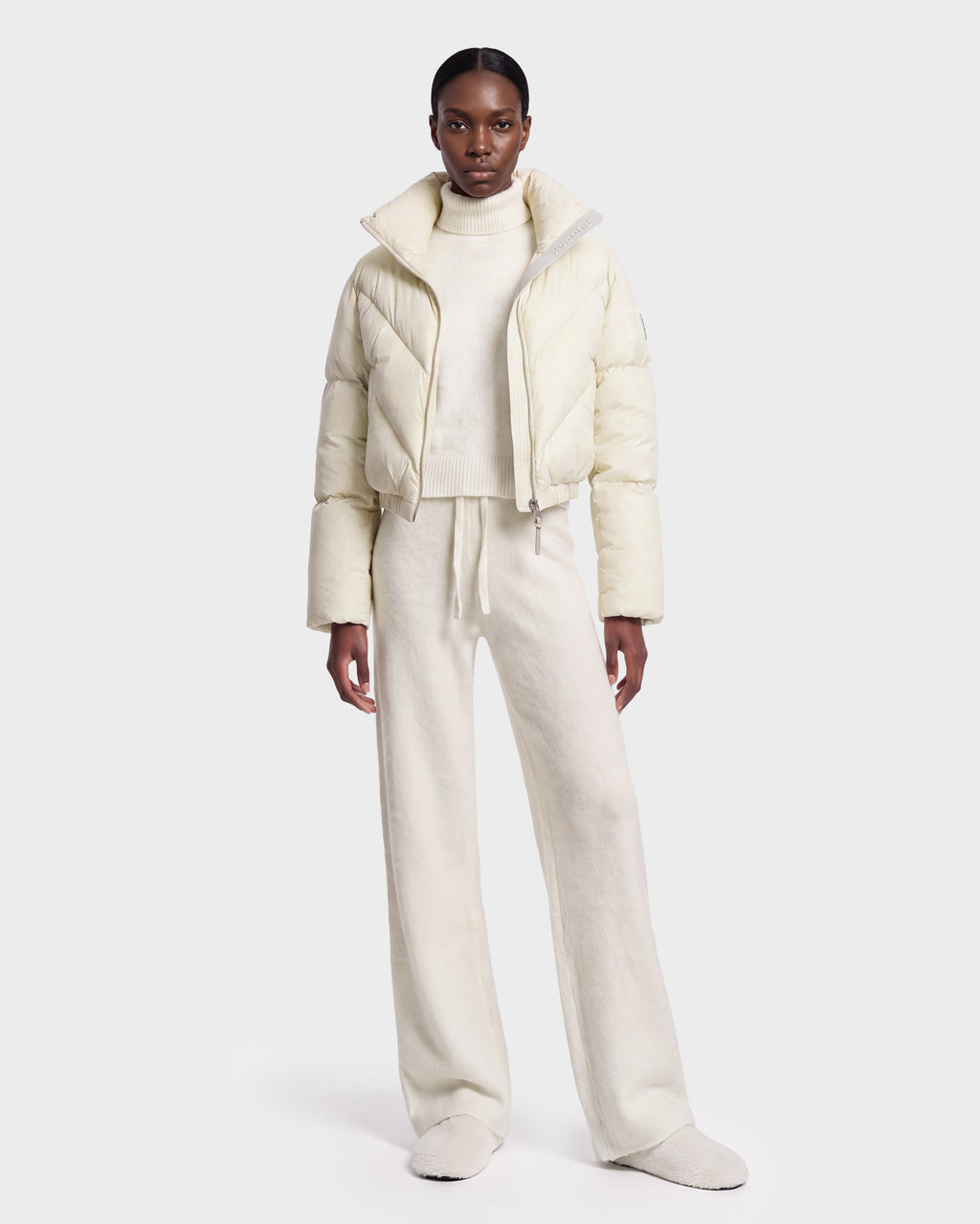 A person stands facing forward against a plain white background, wearing the Rudsak ANSLIE - 8126986 Vanilla off-white cropped puffer jacket filled with RDS-certified down, paired with a turtleneck sweater, wide-leg pants, and slippers.