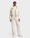 A person stands facing forward against a plain white background, wearing the Rudsak ANSLIE - 8126986 Vanilla off-white cropped puffer jacket filled with RDS-certified down, paired with a turtleneck sweater, wide-leg pants, and slippers.