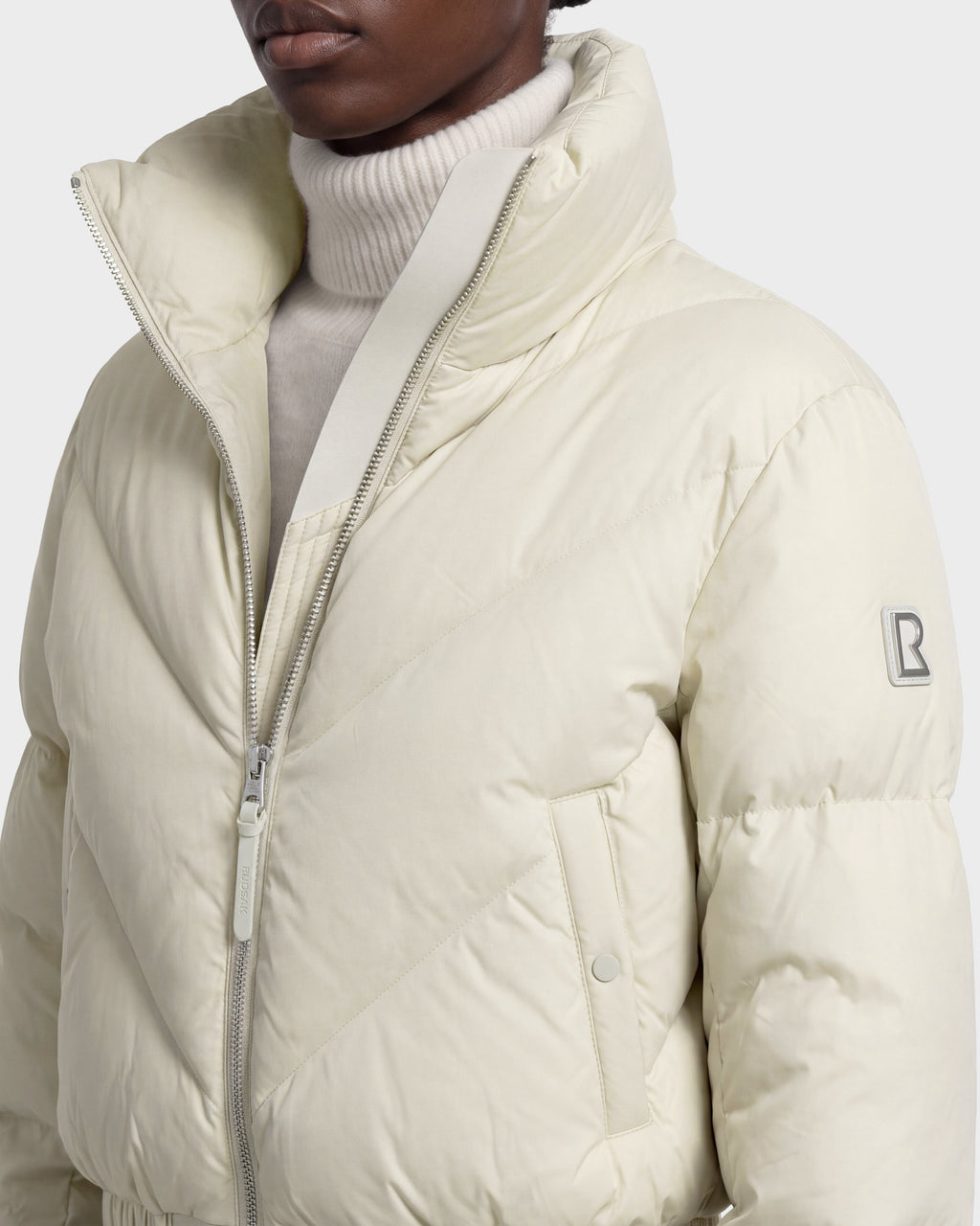 A person is wearing the Rudsak ANSLIE - 8126986 Vanilla, a cream-colored women’s cropped puffer jacket filled with RDS-certified down. This jacket features a zipper closure, high collar, side pockets, and a signature B logo patch on the sleeve, styled over a white turtleneck sweater.