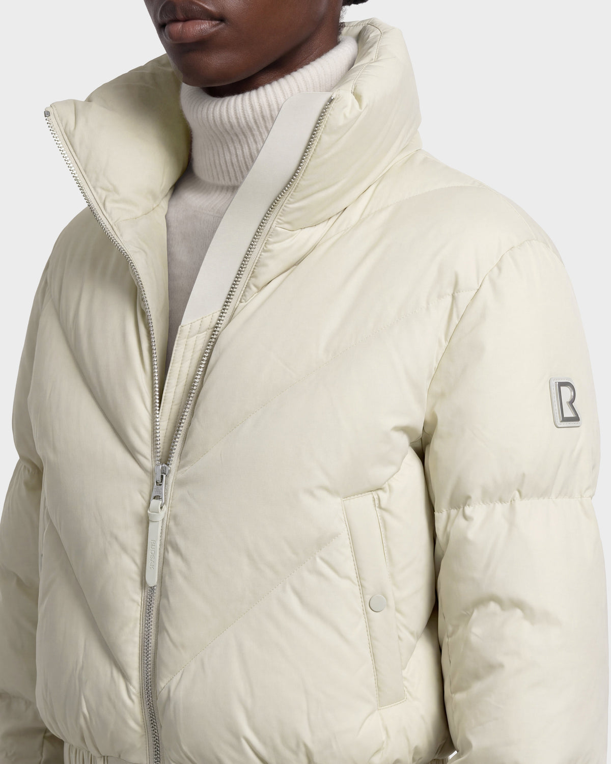 A person is wearing the Rudsak ANSLIE - 8126986 Vanilla, a cream-colored women’s cropped puffer jacket filled with RDS-certified down. This jacket features a zipper closure, high collar, side pockets, and a signature B logo patch on the sleeve, styled over a white turtleneck sweater.