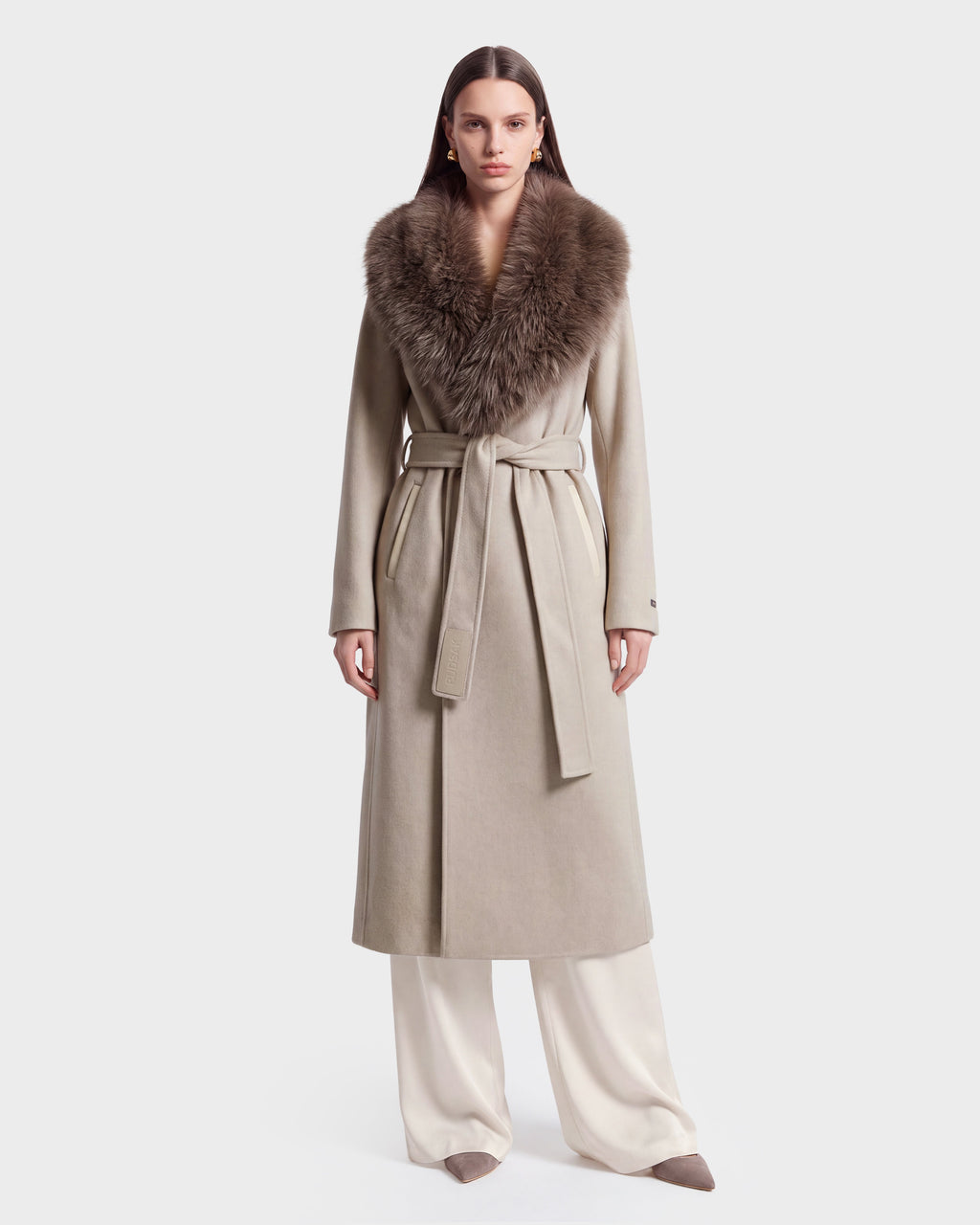 A woman models the Rudsak CINDY - 8126526 Heather Vanilla, a long beige wool coat featuring a wide faux fur collar and matching belt, paired with white pants and beige pointed shoes, standing against a plain white background.
