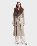 A woman models the Rudsak CINDY - 8126526 Heather Vanilla, a long beige wool coat featuring a wide faux fur collar and matching belt, paired with white pants and beige pointed shoes, standing against a plain white background.