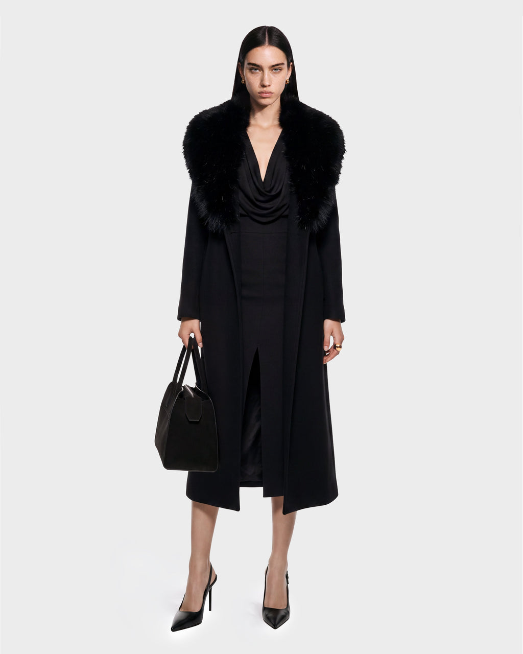 A woman stands facing forward wearing the CINDY - 8126526 Black coat by Rudsak, featuring a fur collar, paired with a black dress with a front slit, black pointed heels, and holding a black handbag. She has straight dark hair and displays a serious expression. This look is available for pre-order and is expected to ship by September 15th.