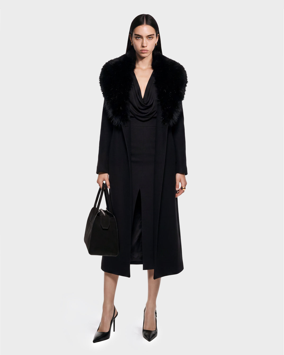 A woman stands facing forward wearing the CINDY - 8126526 Black coat by Rudsak, featuring a fur collar, paired with a black dress with a front slit, black pointed heels, and holding a black handbag. She has straight dark hair and displays a serious expression. This look is available for pre-order and is expected to ship by September 15th.
