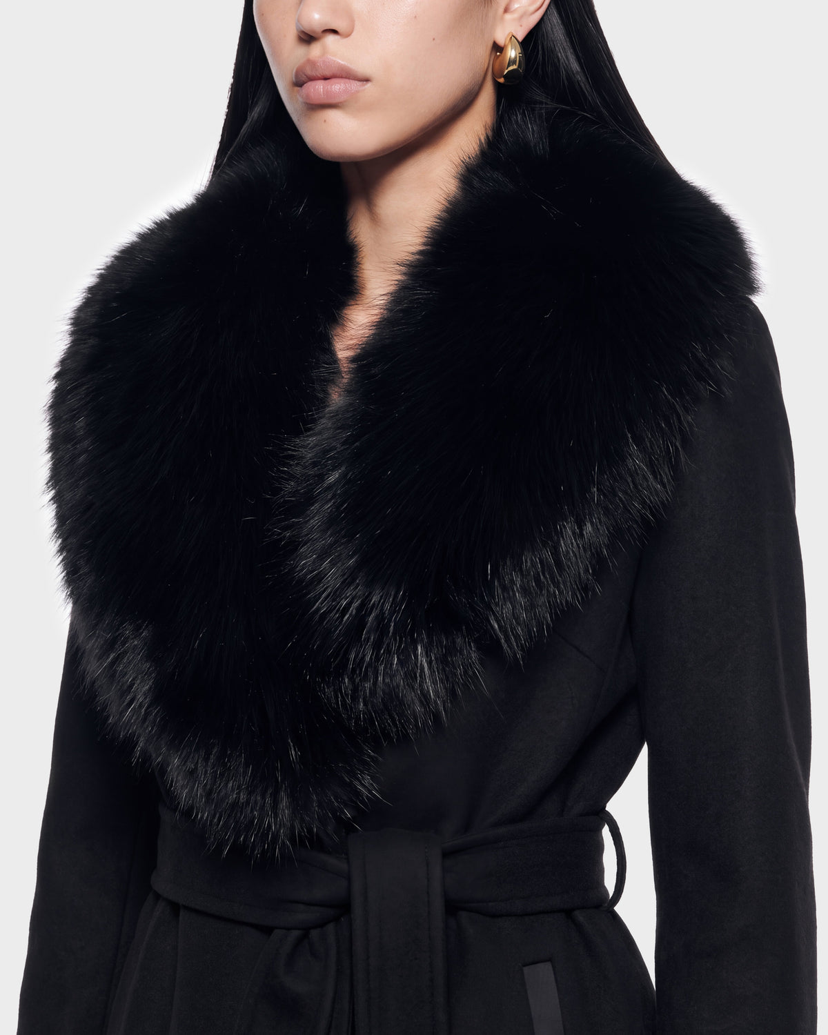 A woman appears from the shoulders up against a plain background, wearing the Rudsak CINDY - 8126526 Black coat with a dramatic, plush black fur collar and gold hoop earrings. This elegant piece is available for pre-order now and is expected to ship by September 15th.