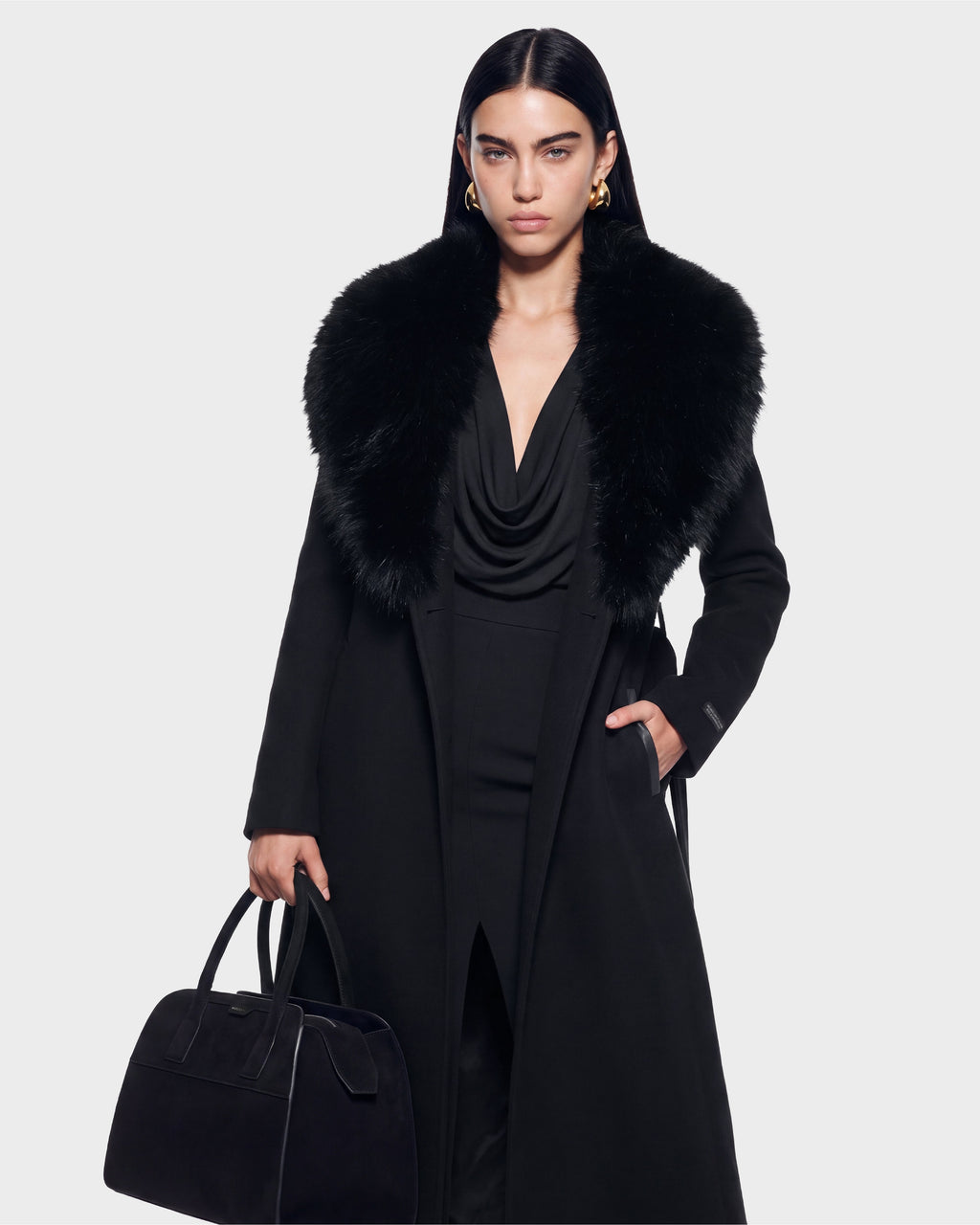 A woman with long dark hair wears the Rudsak CINDY - 8126526 Black coat, featuring a large fur collar, styled with a black dress, gold hoop earrings, and a black handbag against a plain light background. This item is available for pre-order and is expected to ship by September 15th.