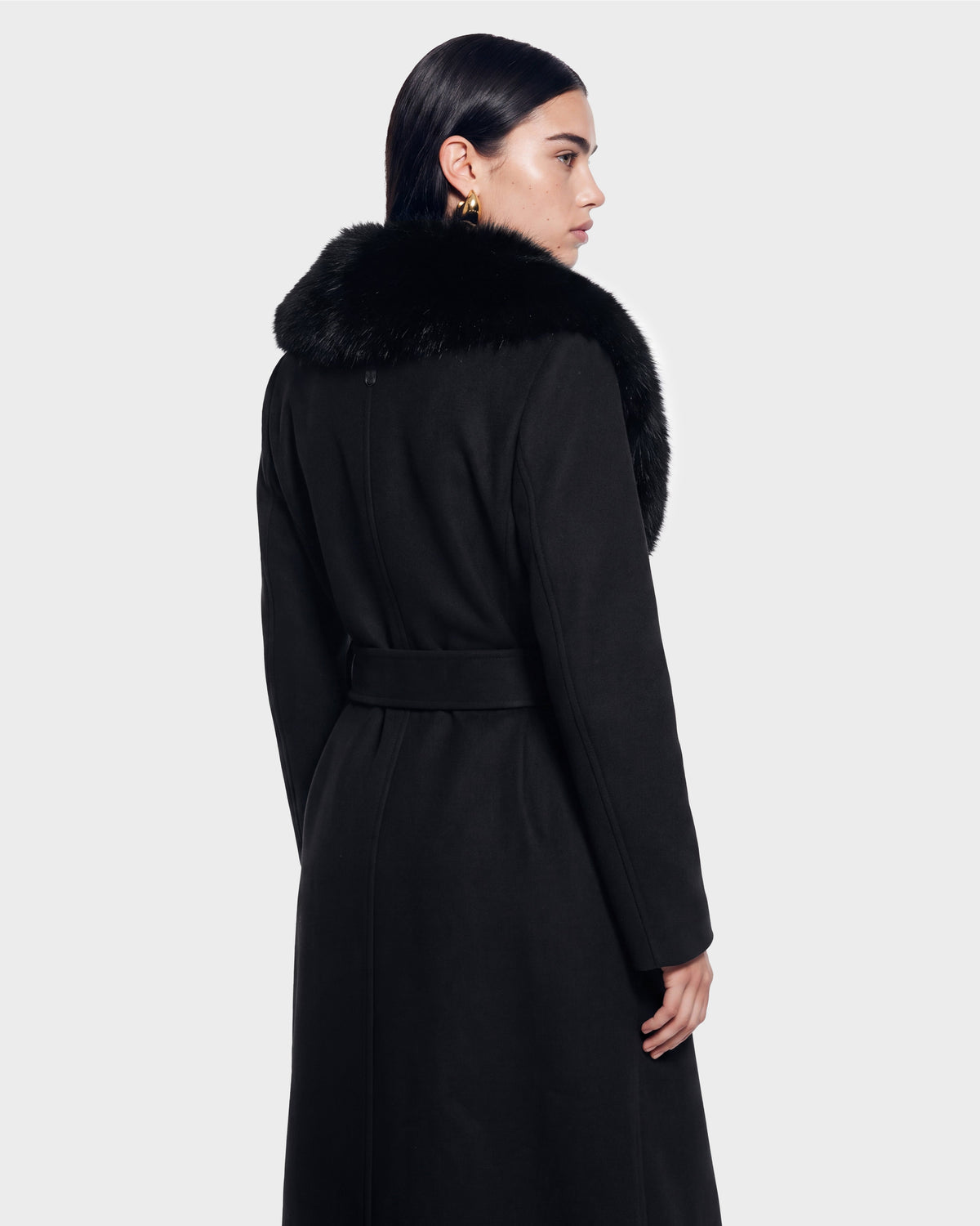 A person with dark hair, gold earrings, and a black belted coat featuring a large black fur collar faces away and looks to the side against a plain light background—this striking look is the CINDY - 8126526 Black by Rudsak, available for pre-order now and expected to ship by September 15th.