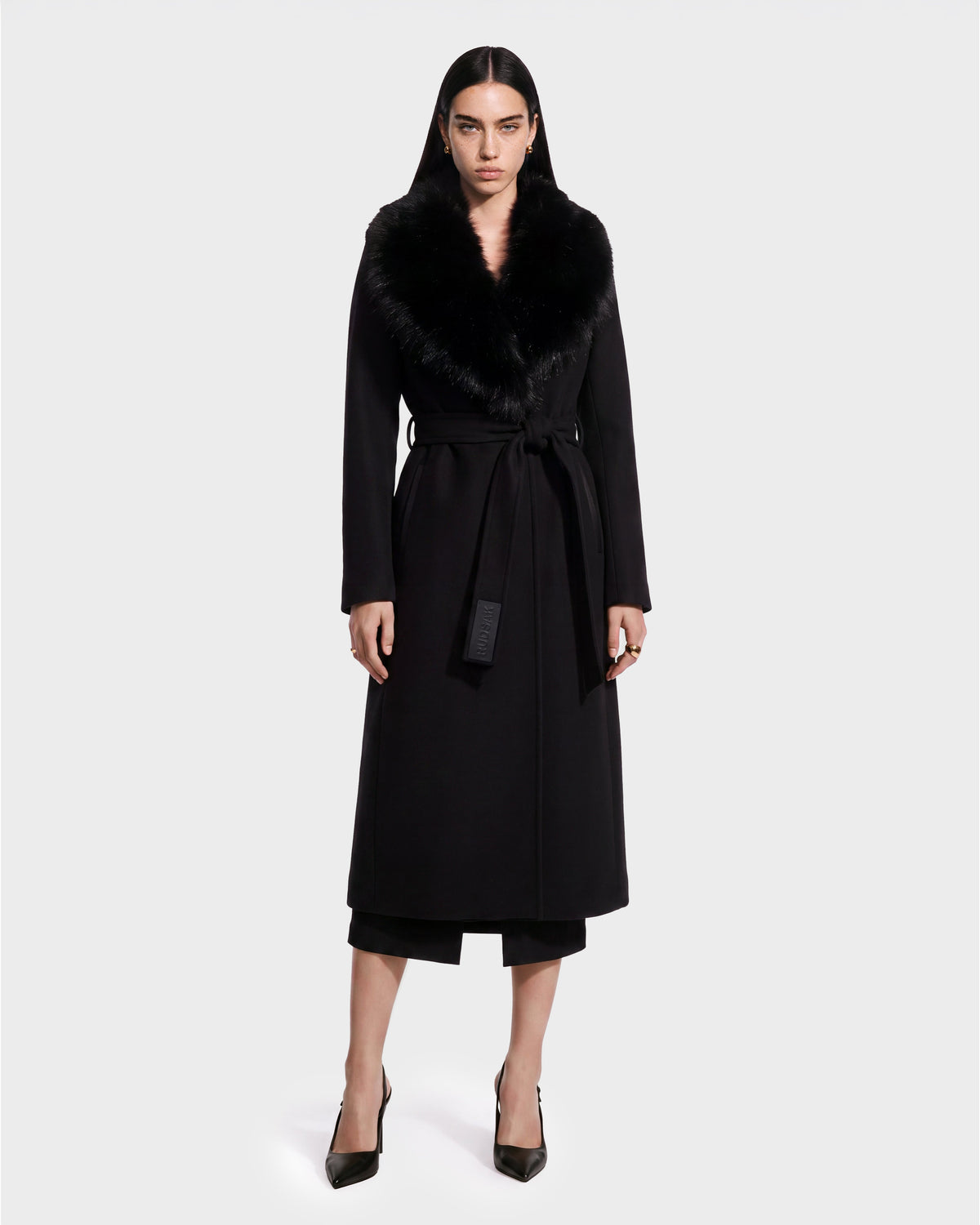 A woman stands facing forward against a plain white background, wearing the Rudsak CINDY - 8126526 Black long coat featuring a wide fur collar, waist belt, black pants, and black heels. This sophisticated piece is available for pre-order now and is expected to ship by September 15th.