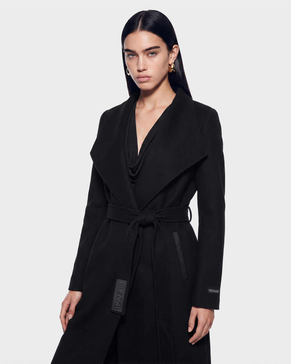 A woman with long dark hair, wearing the Rudsak CINDY - 8126526 Black belted coat with a wide collar, stands against a plain light gray background, looking confidently at the camera. This stylish piece from Rudsak is available for pre-order now and is expected to ship by September 15th.