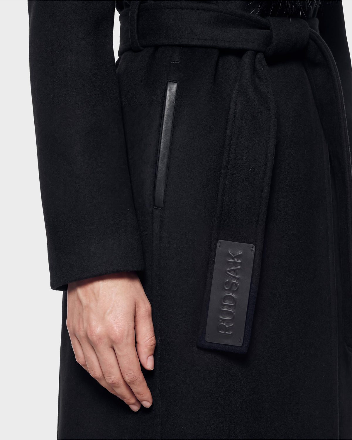 Here is the rewritten sentence incorporating your product data: A close-up of a person’s left hand resting by their side, wearing the CINDY - 8126526 Black coat from Rudsak. The coat features a belted waist with a rectangular RUDSAK tag and a vertical pocket trimmed in leather. Now available for pre-order, this item is expected to ship by September 15th.