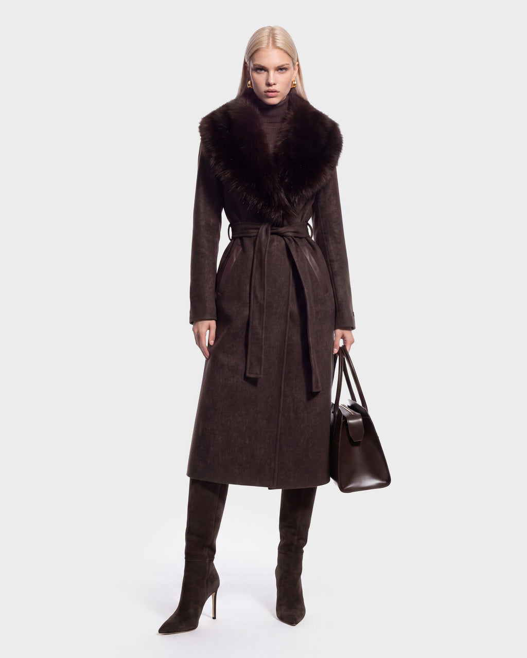 A woman stands facing forward against a plain light background, wearing the Rudsak CINDY - 8126526 Heather Cacao long, dark brown wool coat with a faux fur collar, paired with matching high-heeled boots and carrying a large brown handbag.