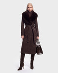 A woman stands facing forward against a plain light background, wearing the Rudsak CINDY - 8126526 Heather Cacao long, dark brown wool coat with a faux fur collar, paired with matching high-heeled boots and carrying a large brown handbag.