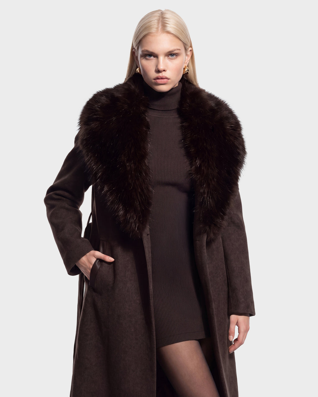 A woman wearing the Rudsak CINDY - 8126526 Heather Cacao long brown wool coat with a large faux fur collar over a ribbed brown dress, standing with one hand in her pocket against a plain background.