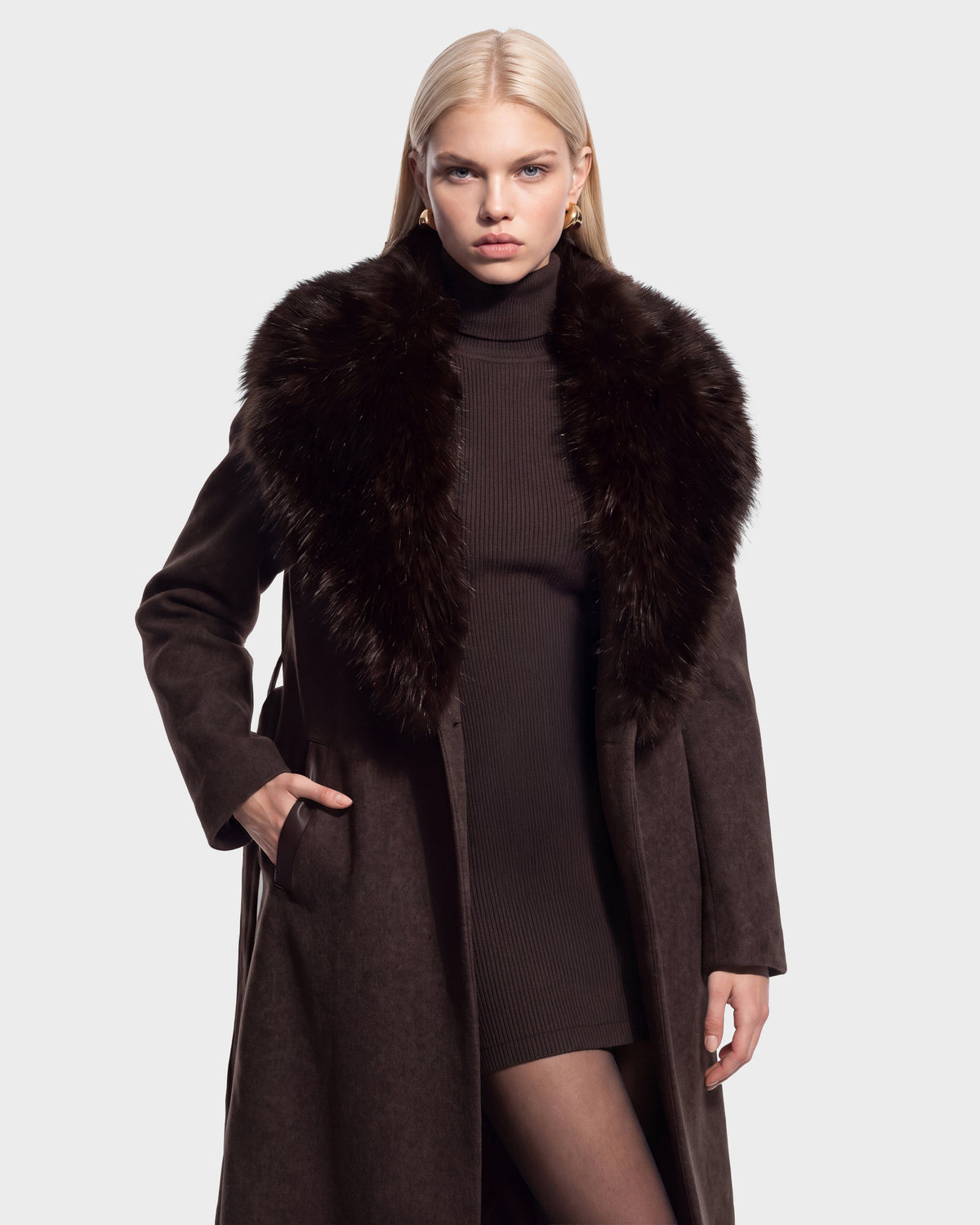 A woman wearing the Rudsak CINDY - 8126526 Heather Cacao long brown wool coat with a large faux fur collar over a ribbed brown dress, standing with one hand in her pocket against a plain background.