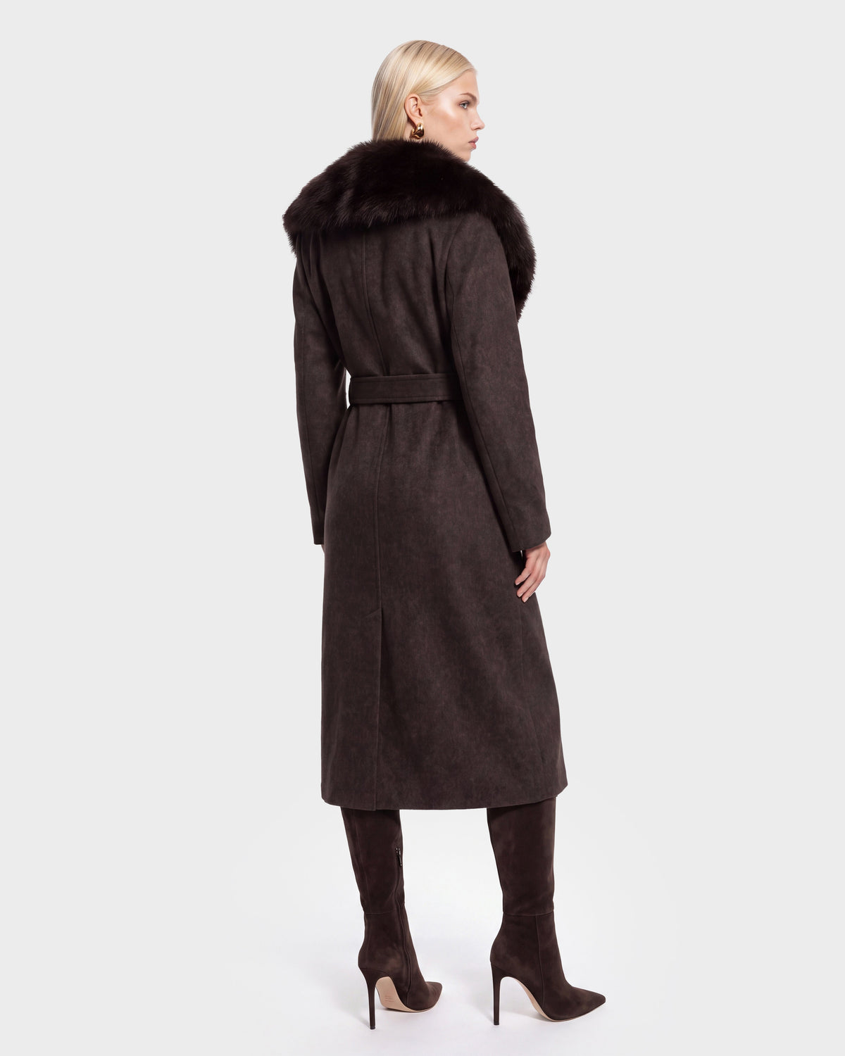 A woman with blonde hair models the Rudsak CINDY - 8126526 Heather Cacao long wool coat, which features a luxurious faux fur collar and is paired with matching tall boots. She stands facing away from the camera against a plain light background.