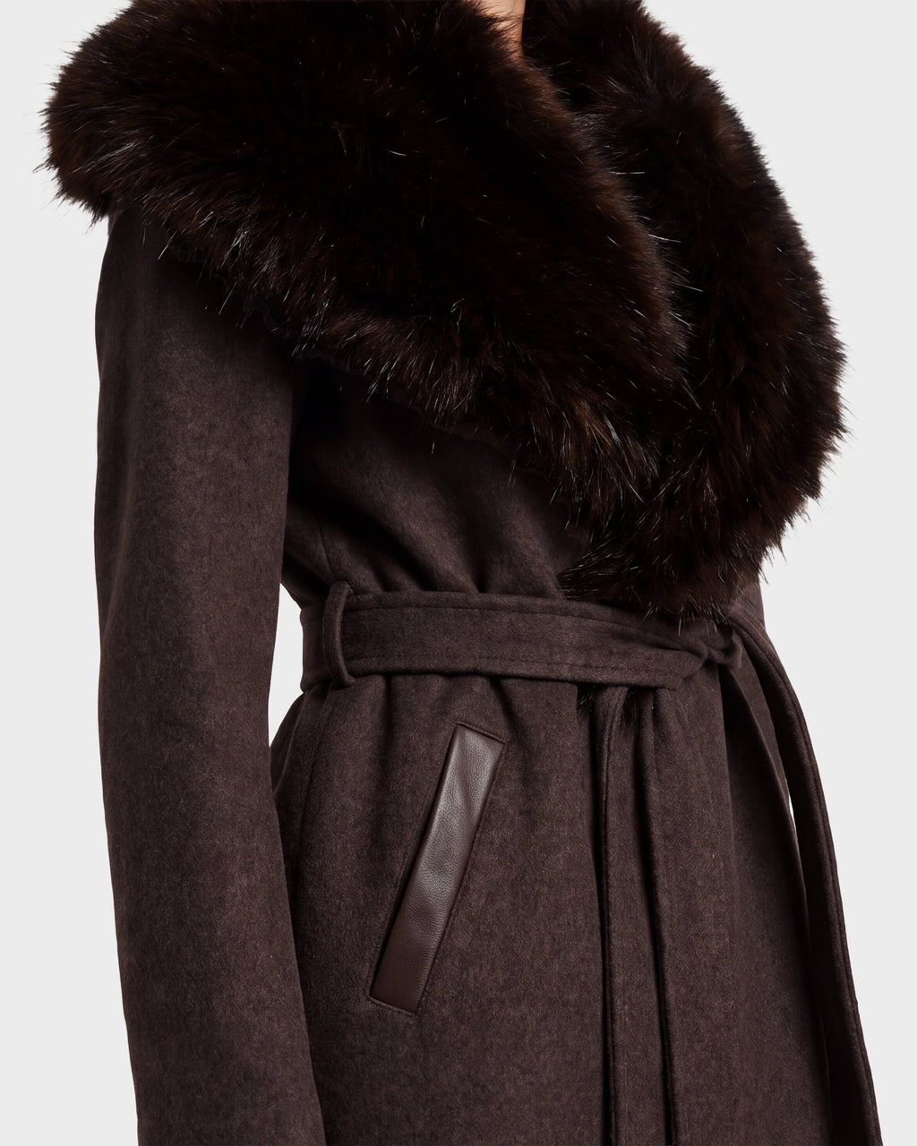 A close-up of a person wearing the Rudsak CINDY - 8126526 Heather Cacao coat, featuring dark brown wool, a large fluffy faux fur collar, leather-trimmed pockets, and a stylish belt.