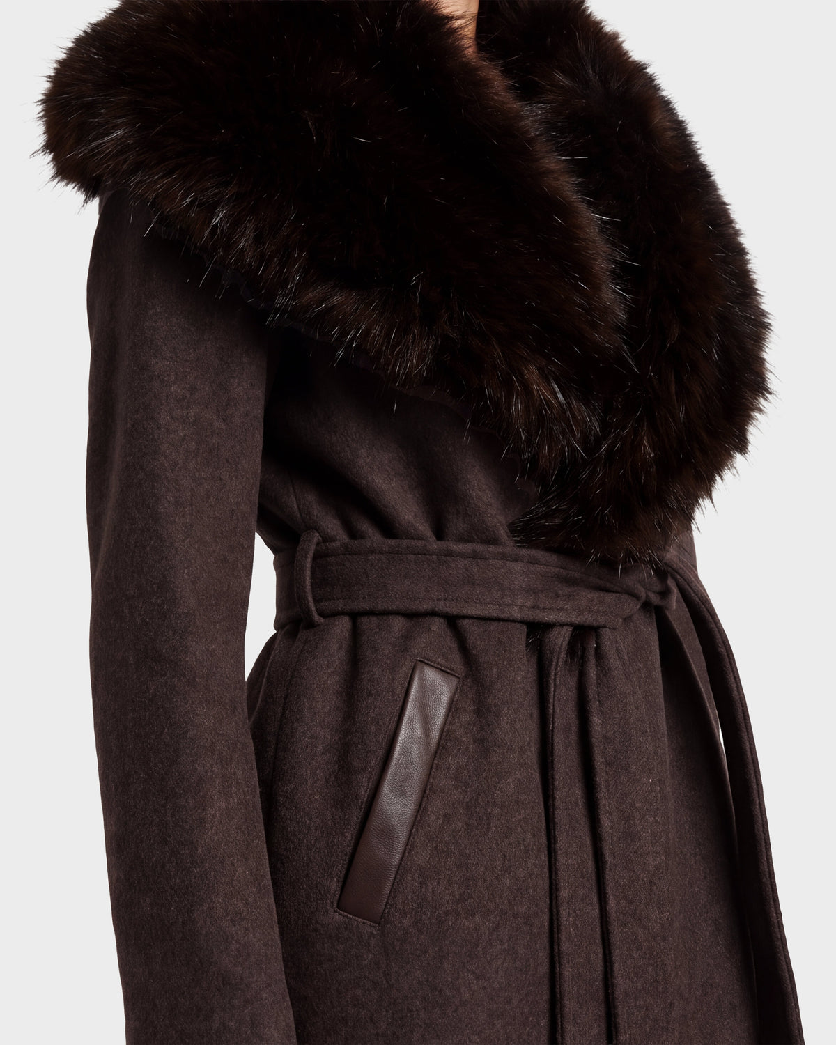 A close-up of a person wearing the Rudsak CINDY - 8126526 Heather Cacao coat, featuring dark brown wool, a large fluffy faux fur collar, leather-trimmed pockets, and a stylish belt.