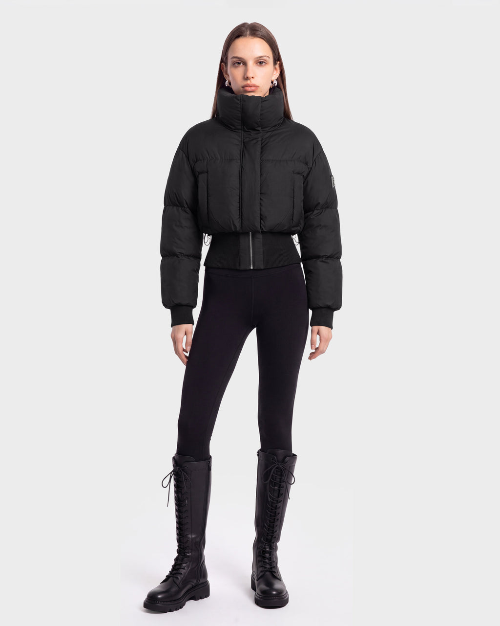 A person stands facing forward, wearing the Rudsak DONYA - 8126516 Black puffer jacket, styled with black leggings and tall black lace-up boots against a plain light gray background. This look is available for pre-order and is expected to ship by September 15th.
