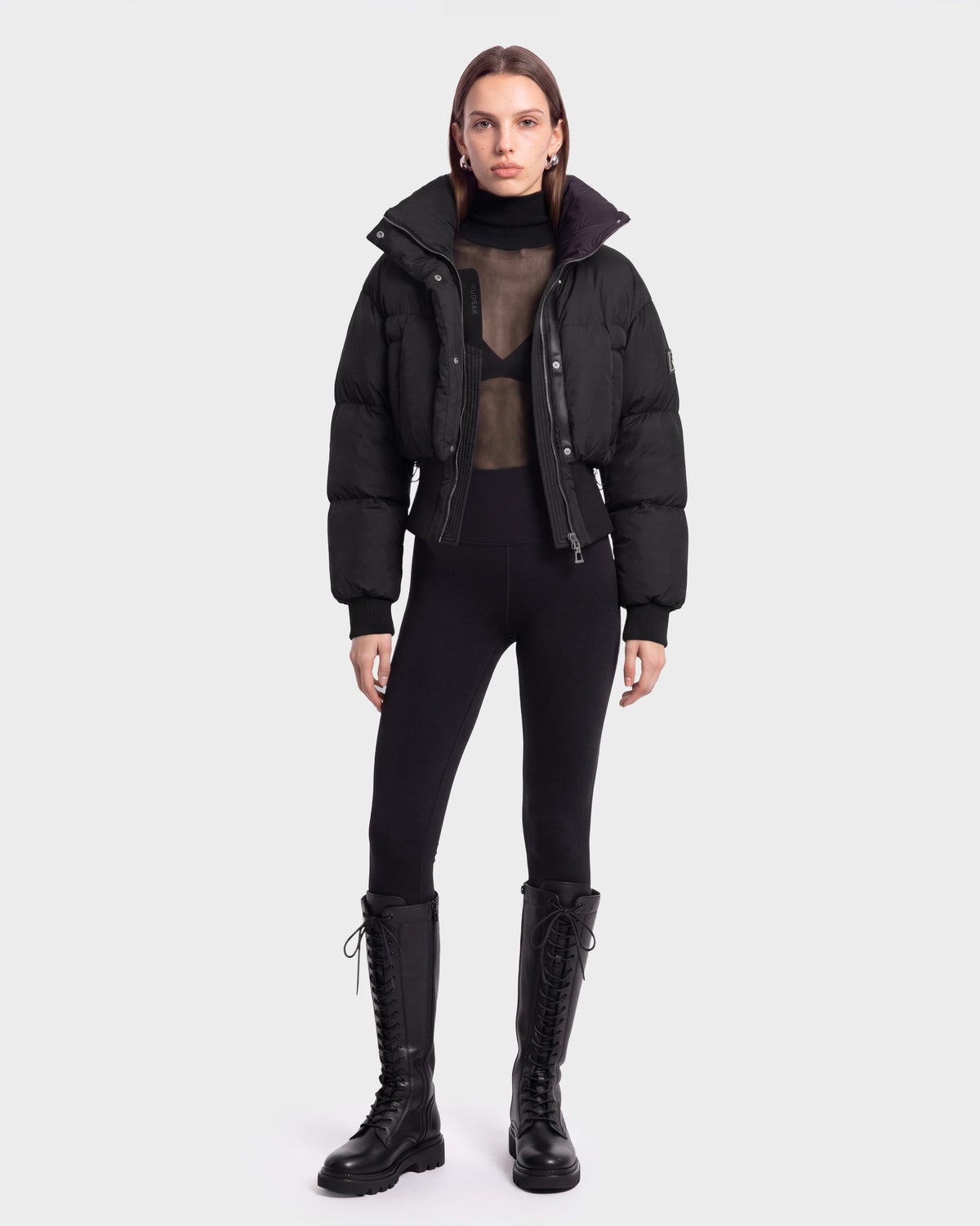 A model stands facing forward against a plain white background, wearing the Rudsak DONYA - 8126516 Black puffer jacket styled with a sheer black top, black leggings, and tall lace-up boots—a look now available for pre-order and expected to ship by September 15th.