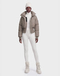 A person wearing the Rudsak DONYA - 8126516 Cement women’s down puffer jacket filled with RDS-certified down, paired with a white knit beanie, a white turtleneck, white leggings, and beige lace-up boots stands against a plain white background.