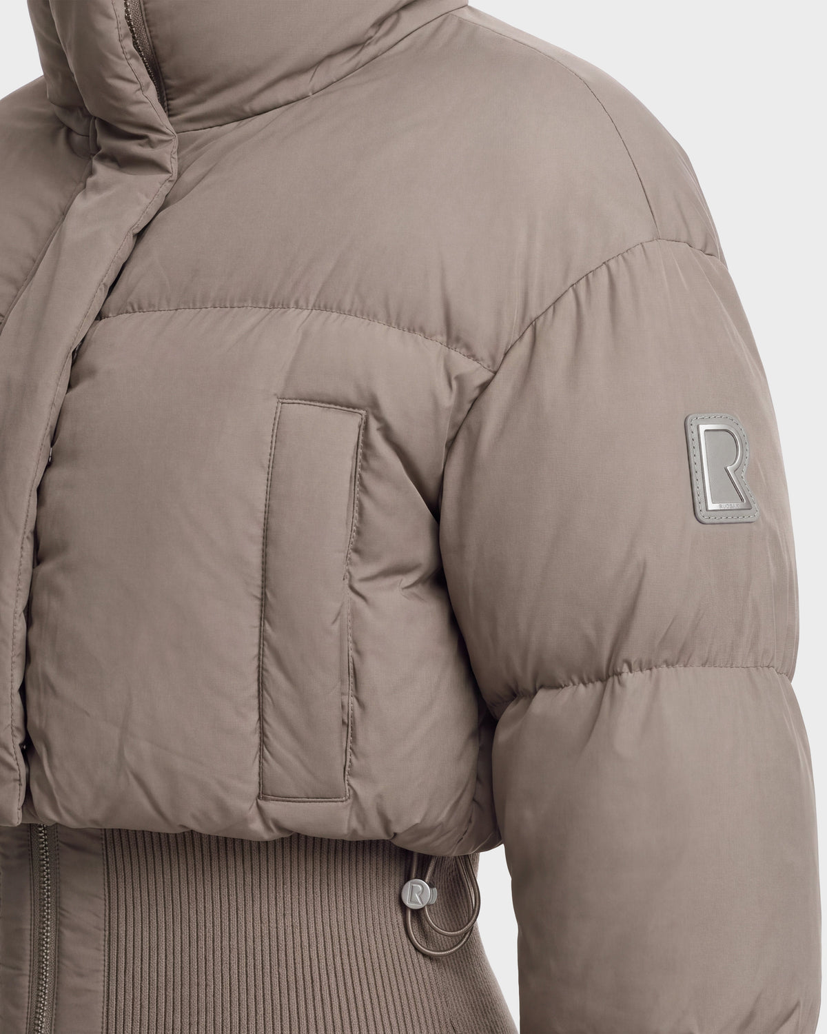 Close-up of the DONYA - 8126516 Cement women’s down puffer jacket by Rudsak in taupe. This winter jacket features a ribbed hem, chest pocket, and an R logo patch on the sleeve. Crafted from matte, soft-textured fabric and filled with RDS-certified down for added warmth, it also offers a high collar for extra protection against the cold.