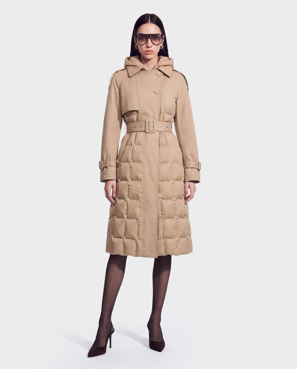 A woman stands against a plain white background wearing the ELVIE - 8126512 Almond quilted down trench coat by Rudsak—belted, tan-hued, and hooded, crafted for superior cold-weather performance with RDS-certified down—paired with oversized sunglasses, sheer black tights, and brown high heels.