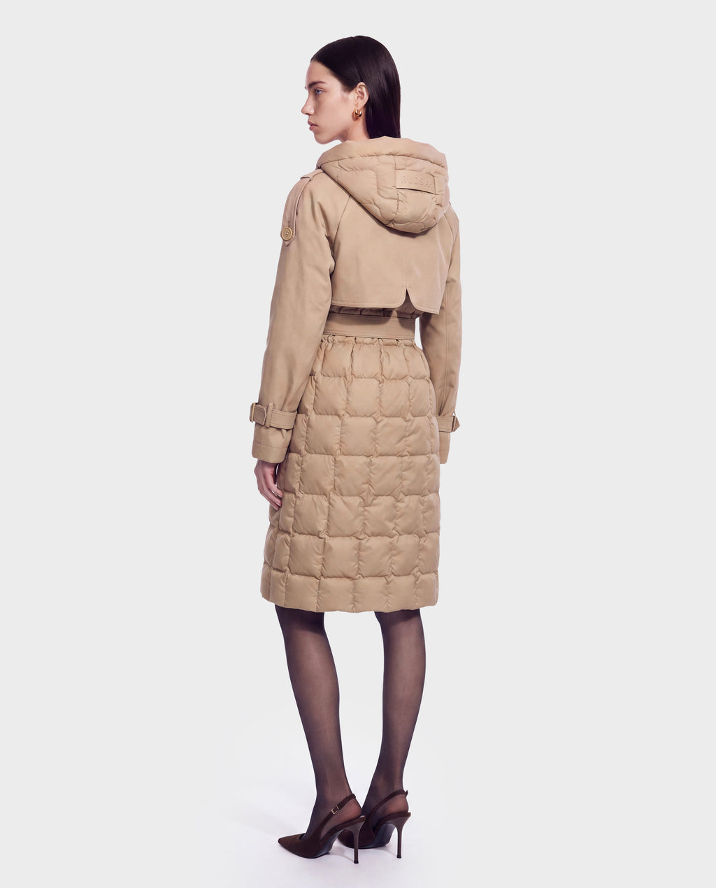 A woman stands facing away, displaying the back view of the Rudsak ELVIE - 8126512 Almond—a tan quilted down trench coat featuring a hood and belt. She pairs her look with dark straight hair, sheer black tights, and brown high-heeled shoes for a chic style that showcases the coat’s cold-weather performance.
