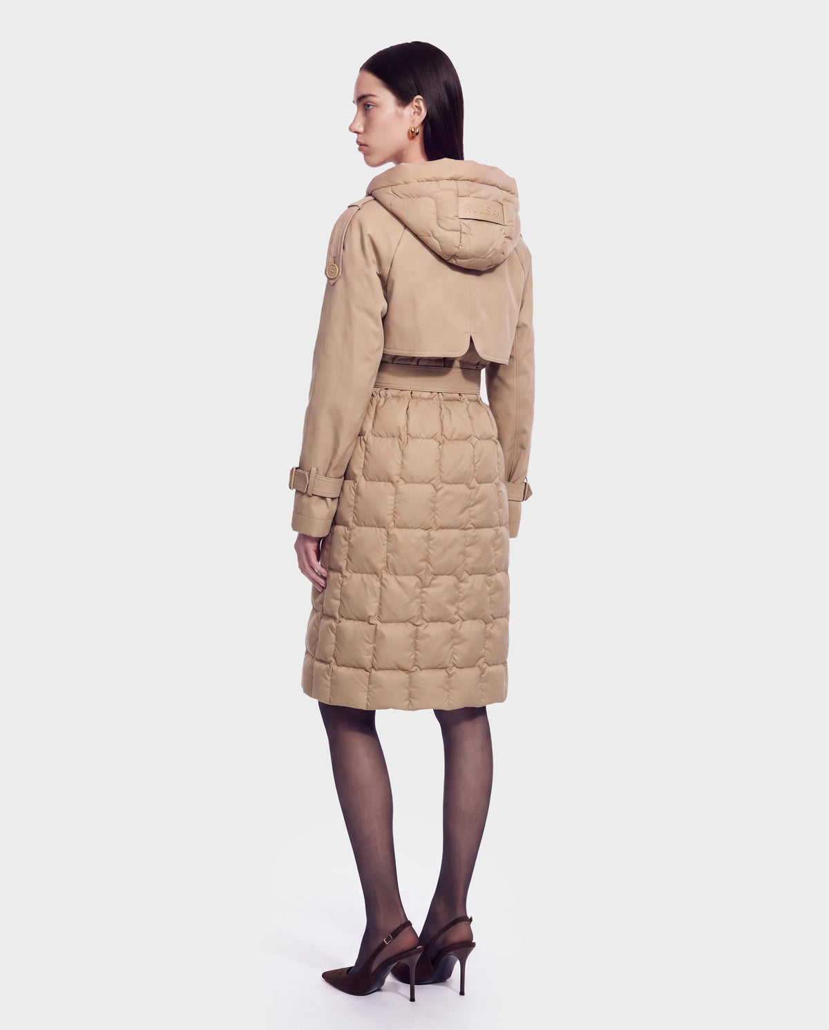 A woman stands facing away, displaying the back view of the Rudsak ELVIE - 8126512 Almond—a tan quilted down trench coat featuring a hood and belt. She pairs her look with dark straight hair, sheer black tights, and brown high-heeled shoes for a chic style that showcases the coat’s cold-weather performance.
