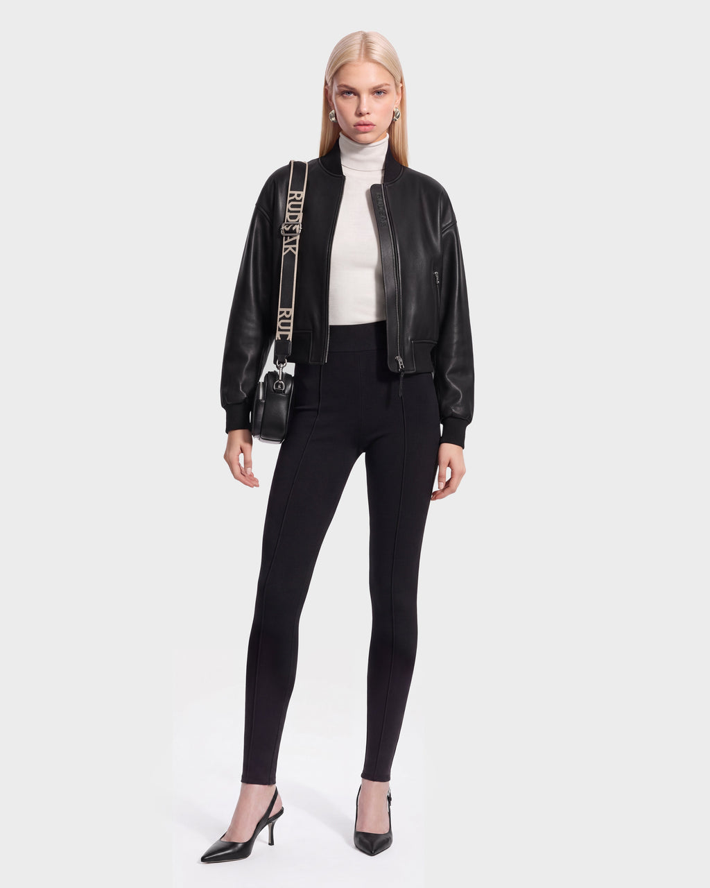 A woman stands against a plain background wearing the EMERSON - 8125041 Black bomber by Rudsak, paired with a white turtleneck, black fitted pants, black heels, and a crossbody bag with a printed strap—effortlessly showcasing contemporary outerwear.
