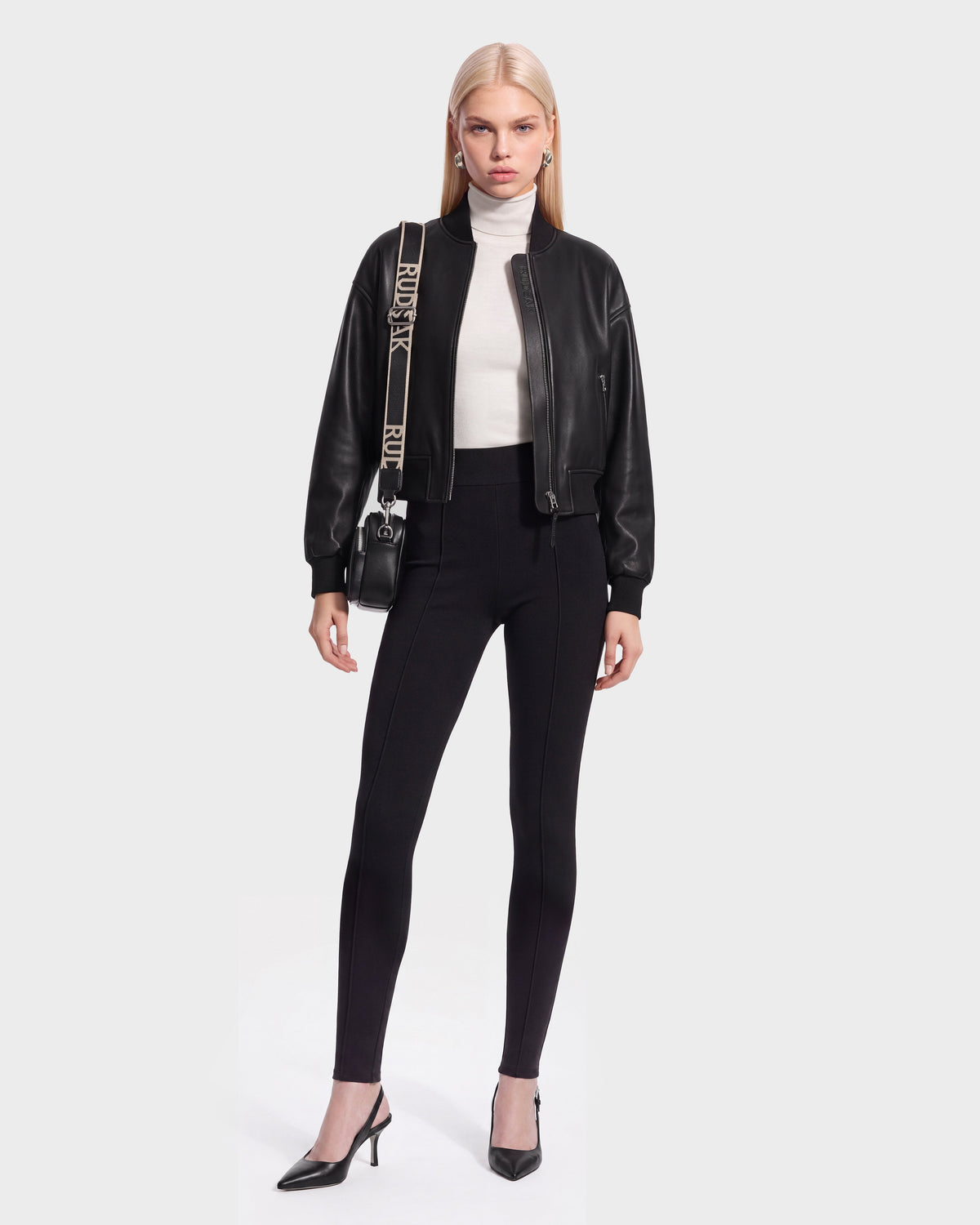 A woman stands against a plain background wearing the EMERSON - 8125041 Black bomber by Rudsak, paired with a white turtleneck, black fitted pants, black heels, and a crossbody bag with a printed strap—effortlessly showcasing contemporary outerwear.