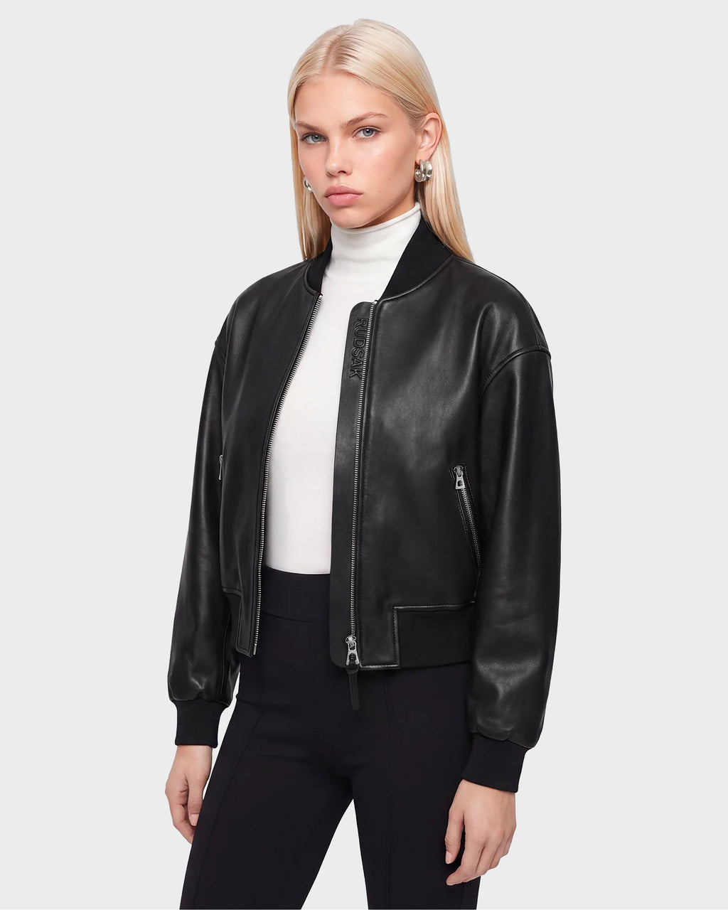 A woman with straight blonde hair models the Rudsak EMERSON - 8125041 Black leather bomber jacket, a standout piece of contemporary outerwear, styled over a white turtleneck and black pants against a simple light background.