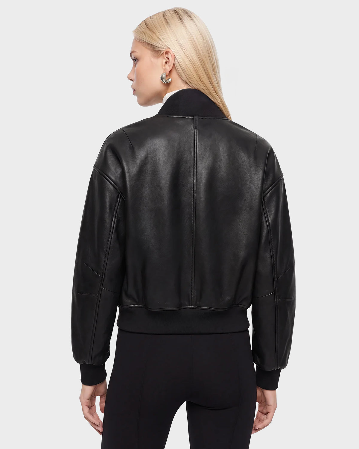 A woman with straight blonde hair wears the Rudsak EMERSON - 8125041 Black leather bomber jacket and black pants, standing with her back to the camera against a plain light gray background—an effortlessly cool take on contemporary outerwear.