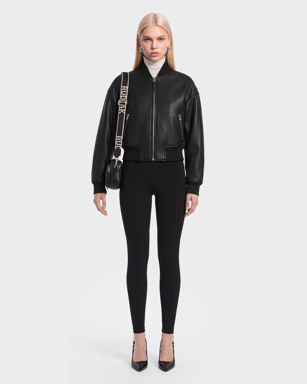 A woman stands facing forward, wearing the EMERSON - 8125041 Black leather bomber jacket by Rudsak, paired with black fitted pants, black heels, and a black crossbody bag with a wide strap. She has straight, long blonde hair and a neutral expression against a plain background.
