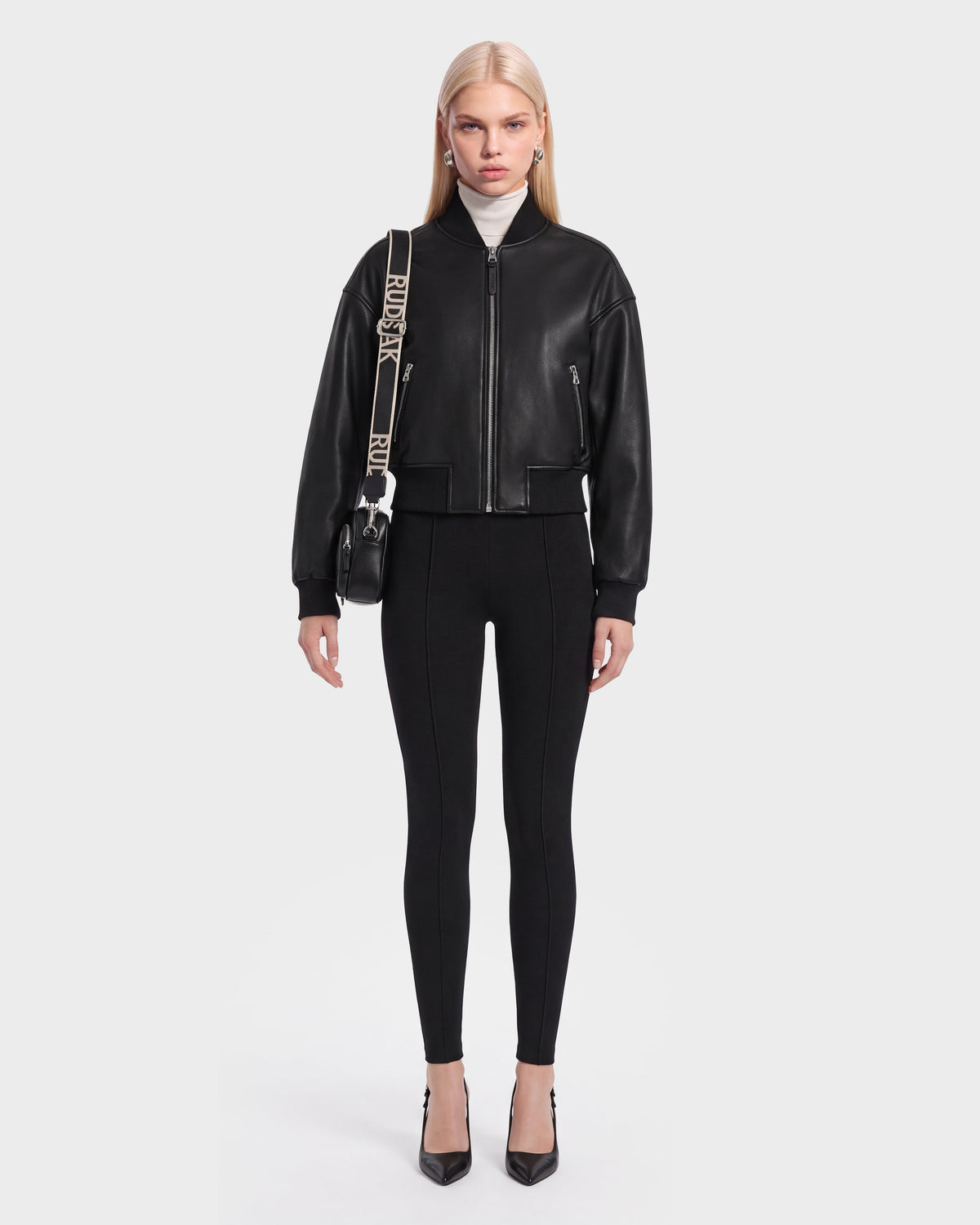 A woman stands facing forward, wearing the EMERSON - 8125041 Black leather bomber jacket by Rudsak, paired with black fitted pants, black heels, and a black crossbody bag with a wide strap. She has straight, long blonde hair and a neutral expression against a plain background.