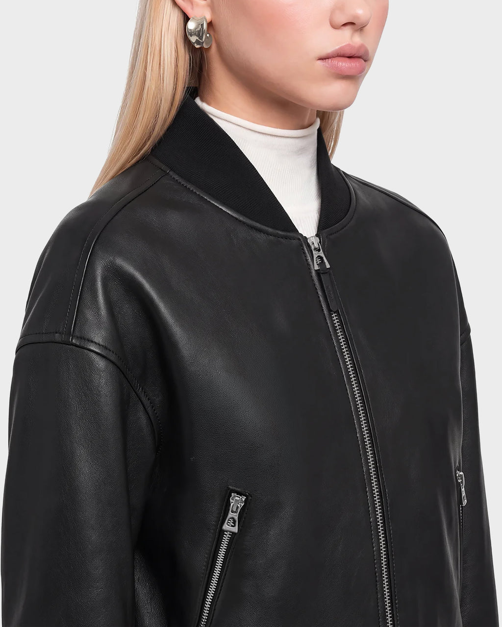 A woman models the Rudsak EMERSON - 8125041 Black leather bomber jacket, featuring silver zippers, styled over a white turtleneck and paired with silver hoop earrings. Her blond hair is worn down and parted to the side, set against a plain light gray background that accentuates her modern, chic outerwear look.