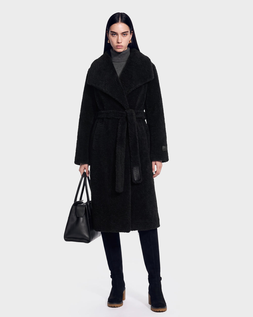 A woman wearing the FRANCIE - 8126873 Black long belted coat by Rudsak, paired with a gray turtleneck and black boots, holds a black handbag. She stands facing forward against a plain white background. This stylish look is available for pre-order and is expected to ship by September 15th.