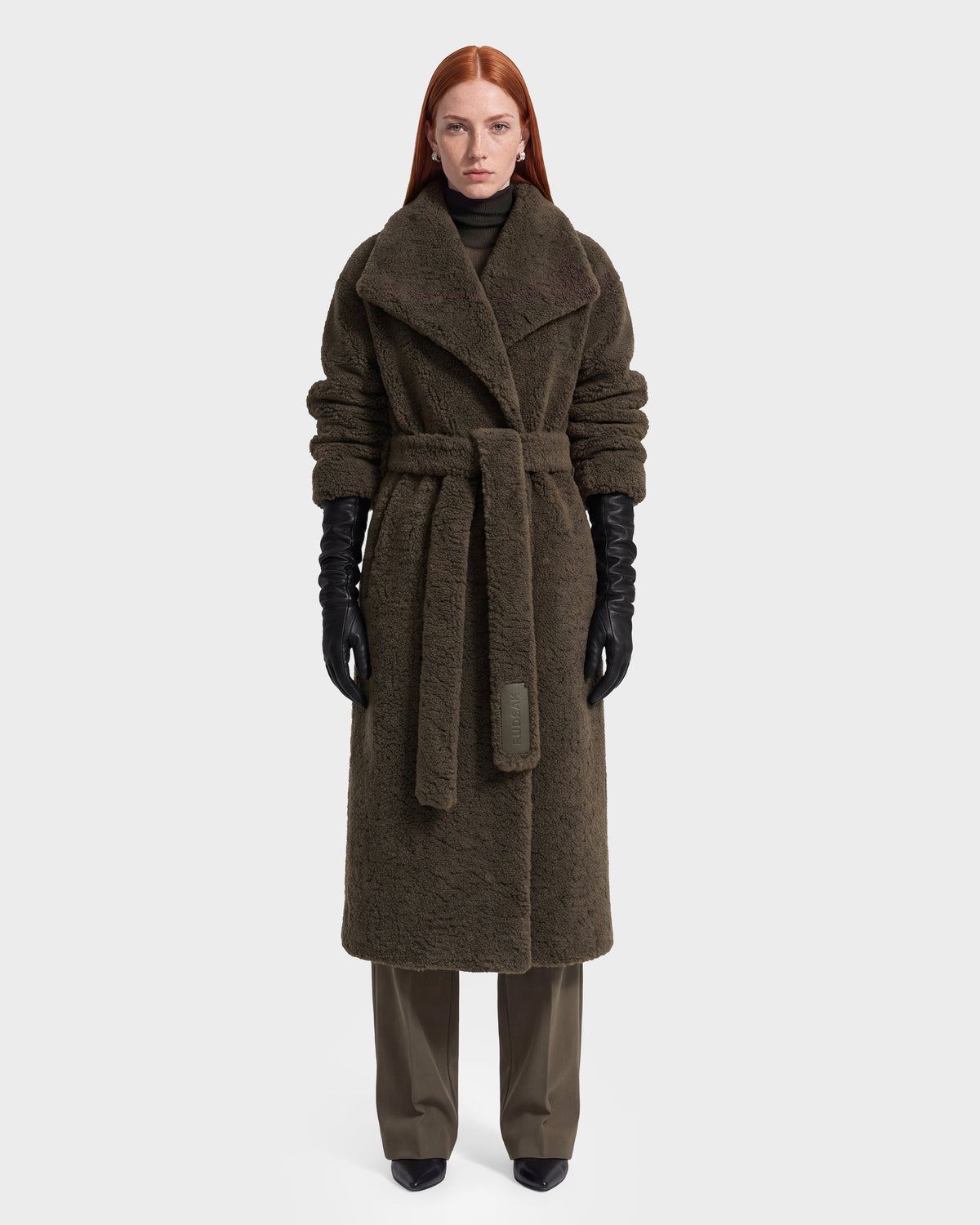 A woman with long red hair models the Rudsak FRANCIE - 8126873 Olive, a long, textured dark brown belted coat, layered over dark pants with black gloves and shoes. Photographed against a plain white background, this piece is available now for pre-order and expected to ship by September 15th.
